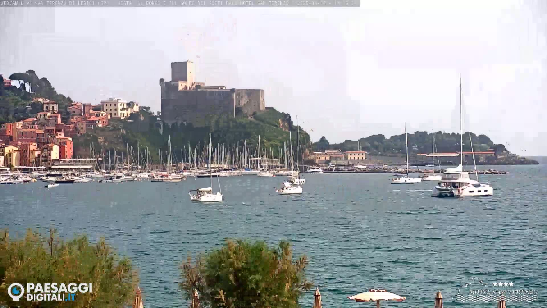 A harbor filled with sailboats is seen in front of a hilltop town and castle under a hazy sky.
