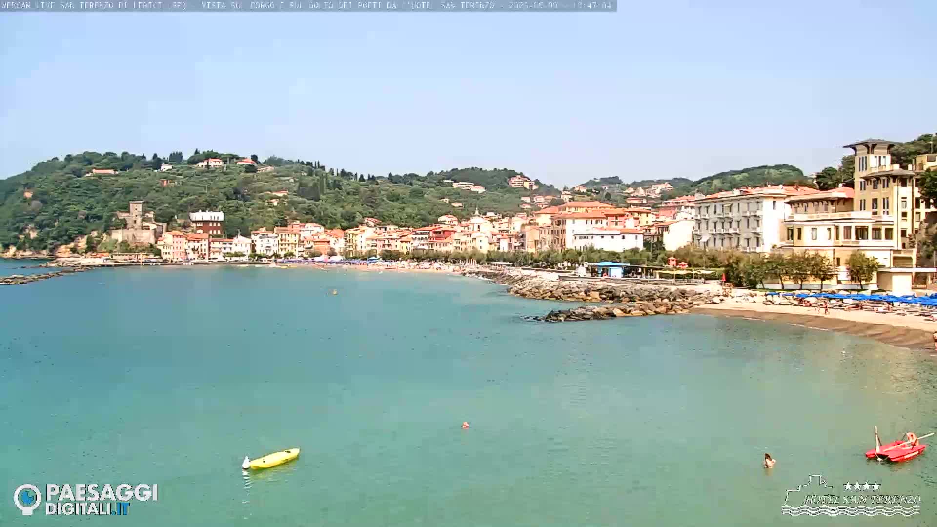 A calm, clear day at a beach town with a sandy beach, clear turquoise water, and buildings on a hillside.