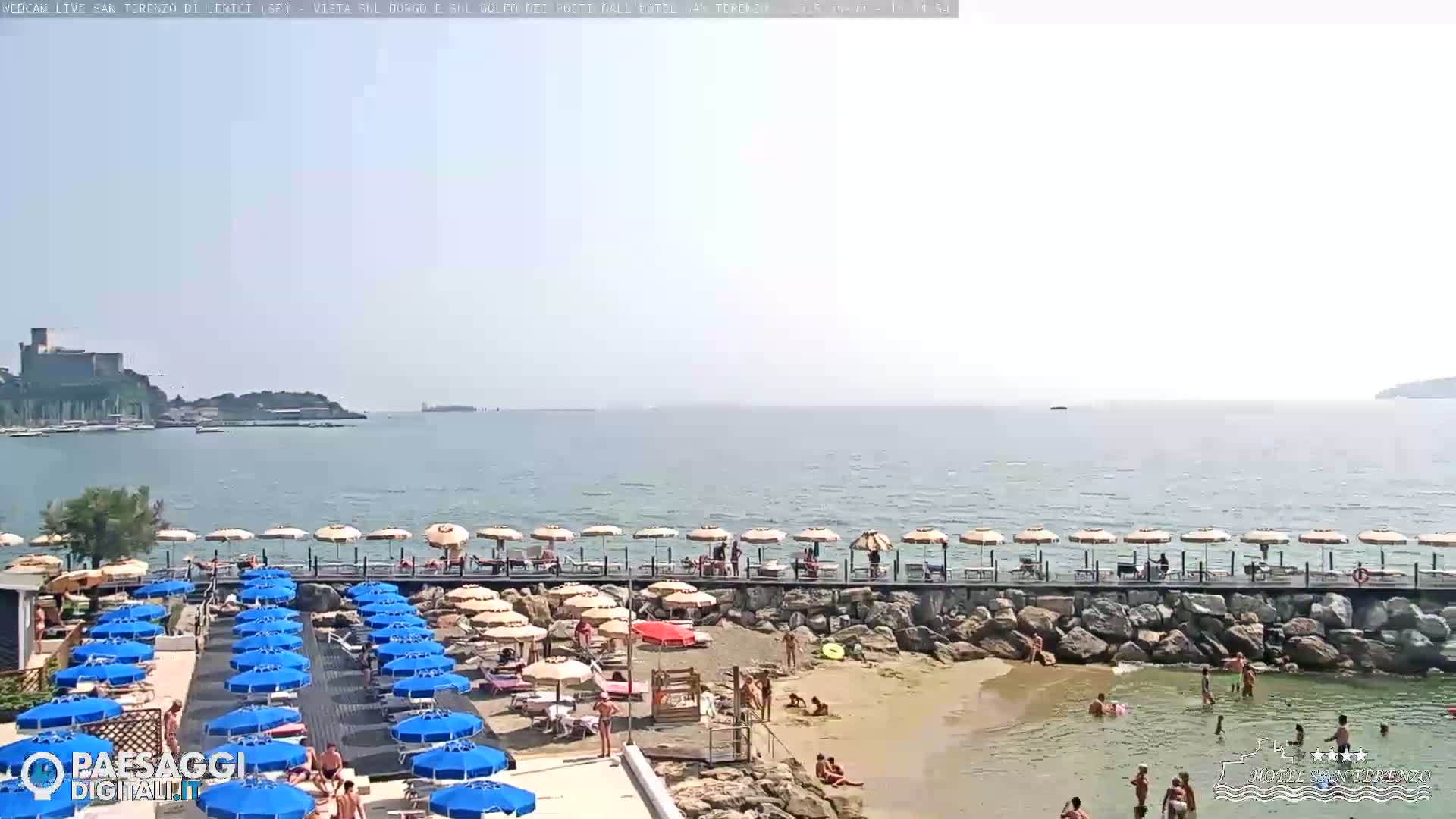 A sandy beach with numerous blue and beige umbrellas, a rocky shoreline, and calm ocean waters under a hazy sky.