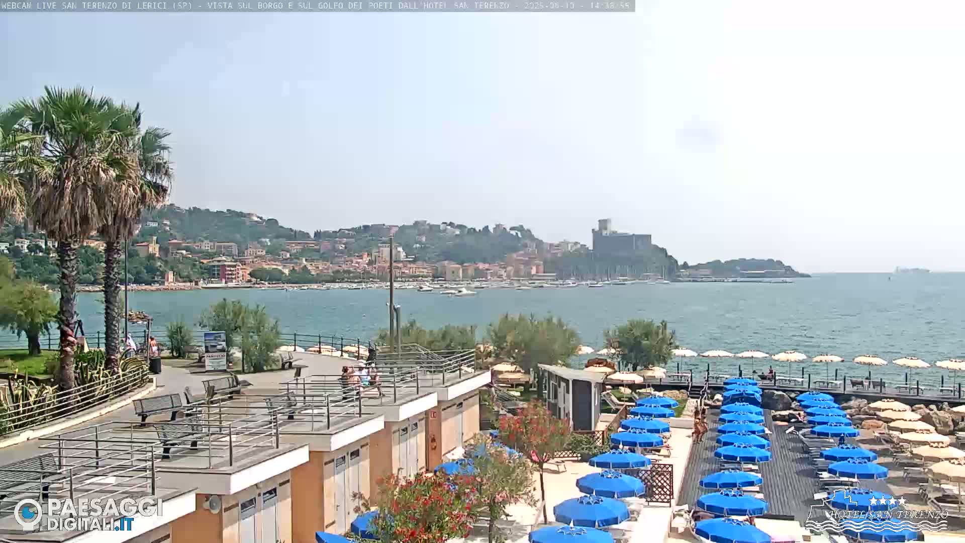 A seaside town with a calm bay, many blue beach umbrellas, and palm trees on a sunny day.