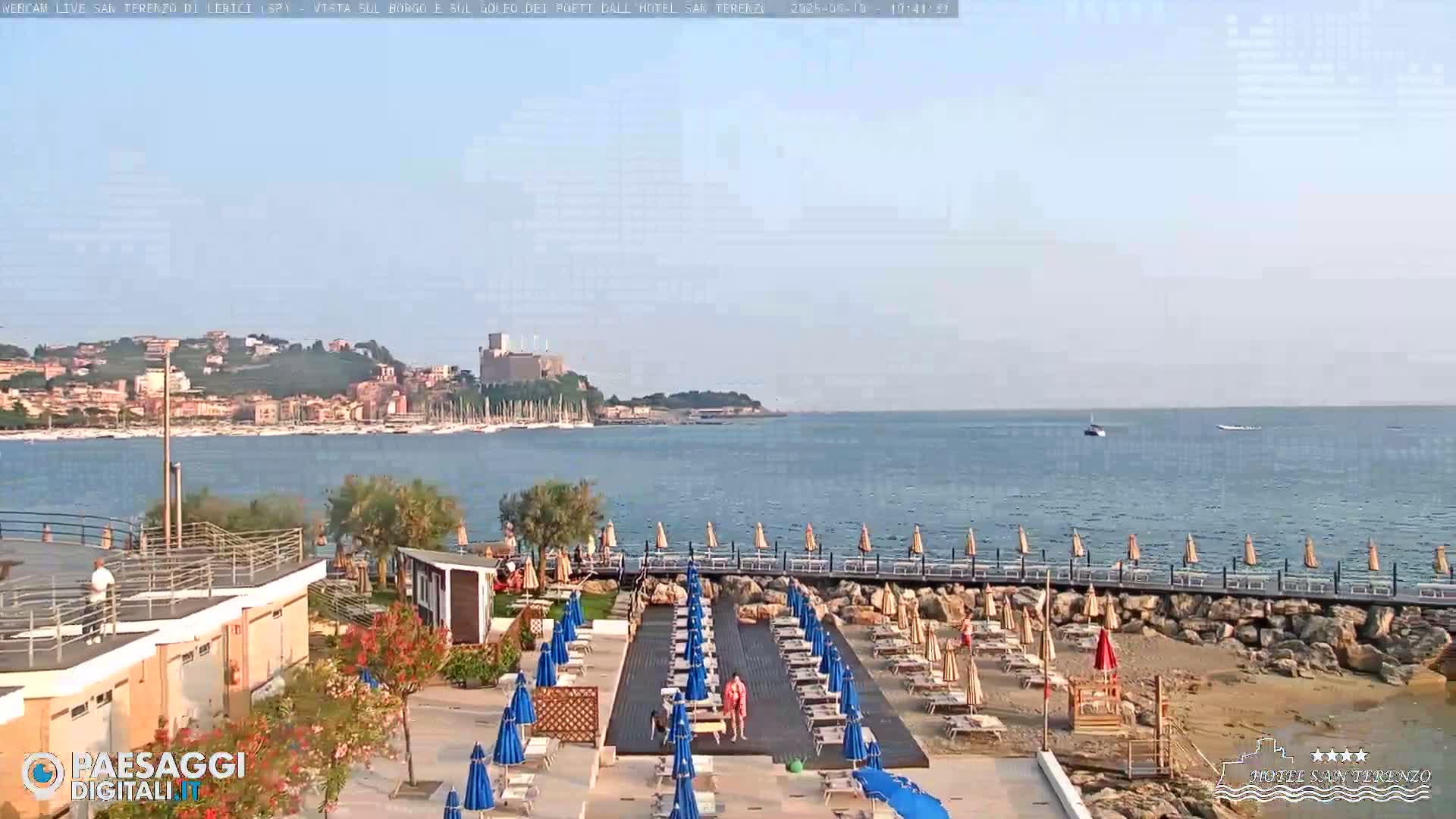 A beach with blue and tan umbrellas, a wooden deck, and a rocky shoreline overlooks a calm bay with boats and a town in the distance under a clear sky.