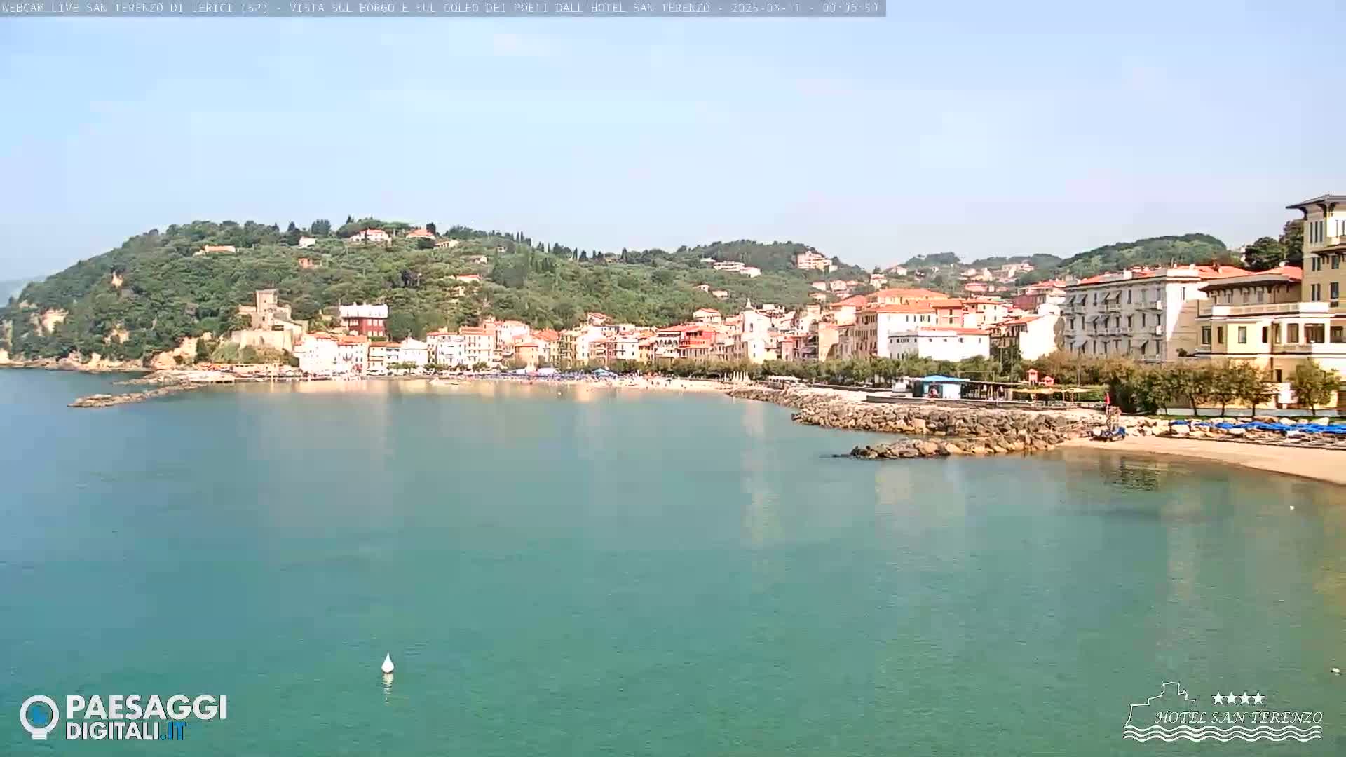 A picturesque coastal town with a sandy beach and calm, clear water sits nestled against a verdant hillside under a sunny sky.