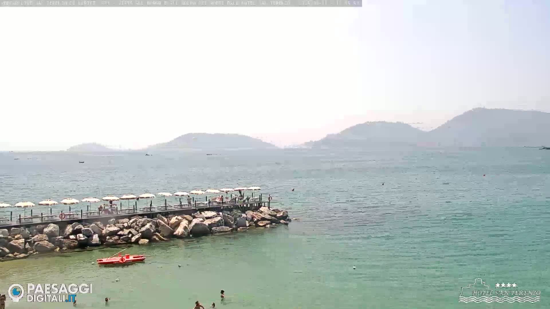 A hazy, sunny day shows a calm sea with a rocky jetty extending into the water, featuring several umbrellas and people, with distant hills visible in the background.