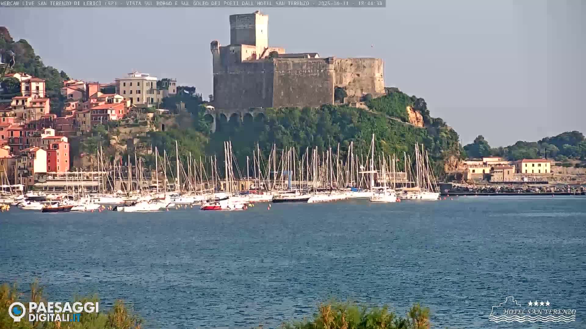 A stone castle sits atop a hill overlooking a calm harbor filled with sailboats and a colorful village.