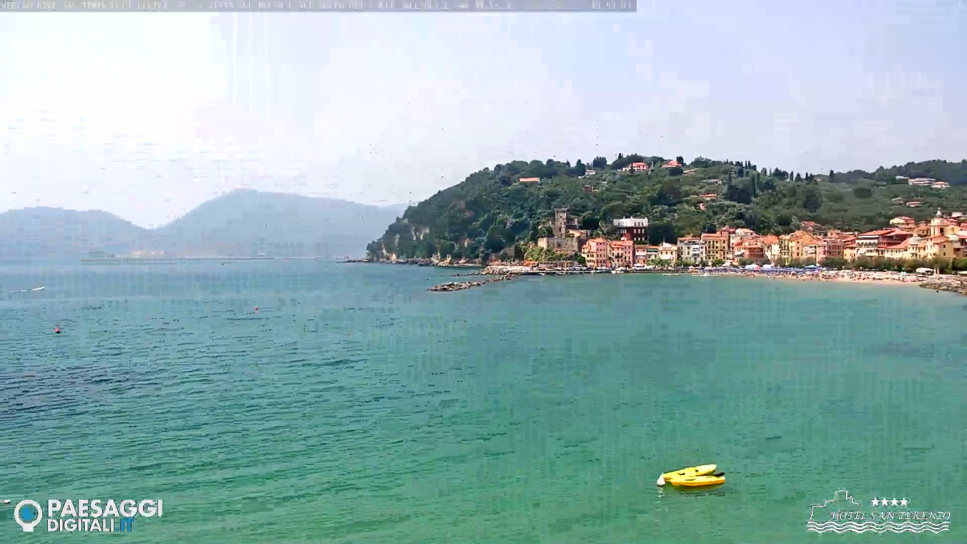 A picturesque coastal town nestled on a verdant hillside, overlooking a calm, teal sea under a mostly clear sky.