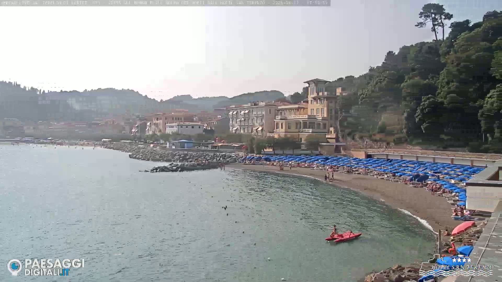 A sandy beach with numerous blue umbrellas and a few people is situated along a calm body of water, backed by buildings and lush green hills under a hazy sky.
