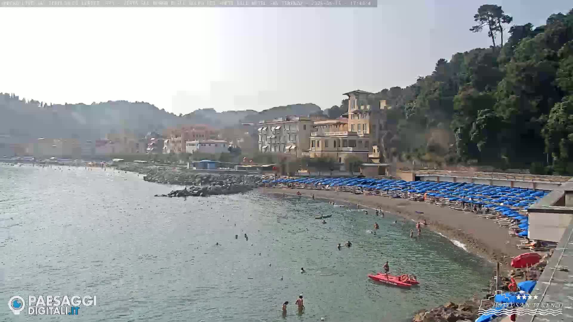 A partly sunny day at a beach with calm water, showing a sandy shore lined with blue umbrellas and beachgoers, backed by buildings and lush green hills.