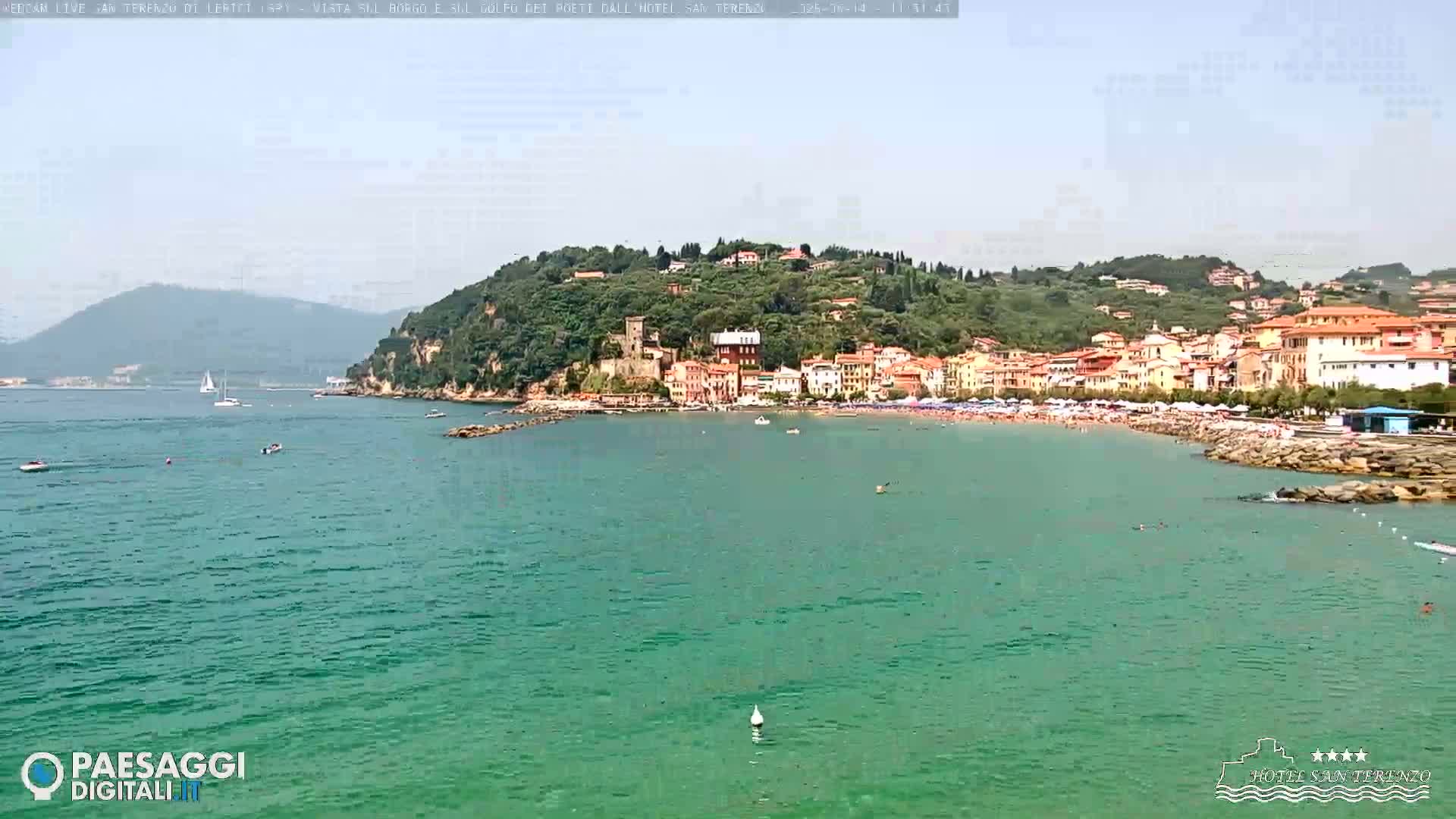 A coastal town with a sandy beach and turquoise water is nestled against a lush green hill under a clear sky.
