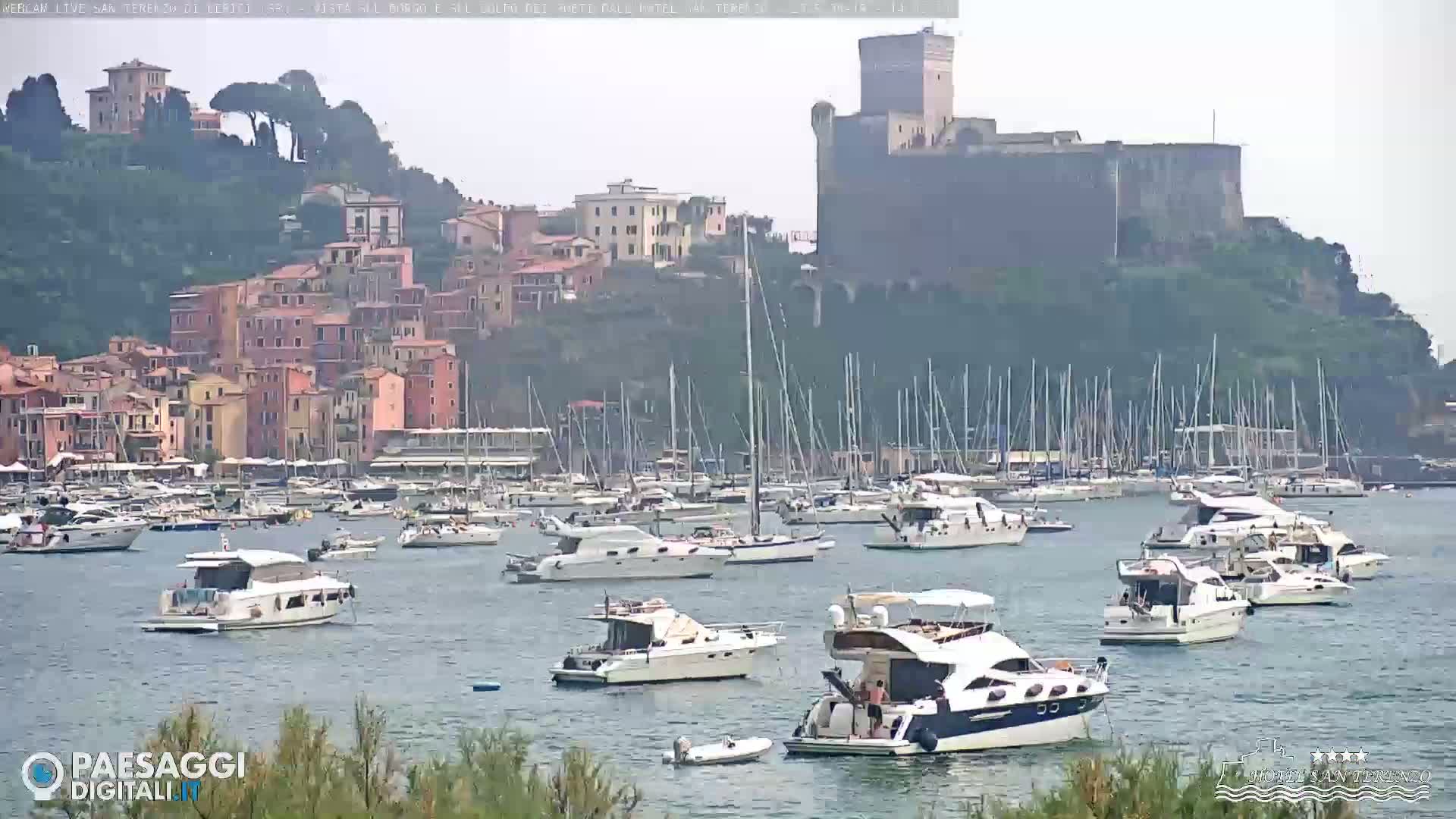 A harbor filled with numerous boats and yachts is situated below a hill topped with a stone castle and a village of closely clustered buildings, all under an overcast sky.