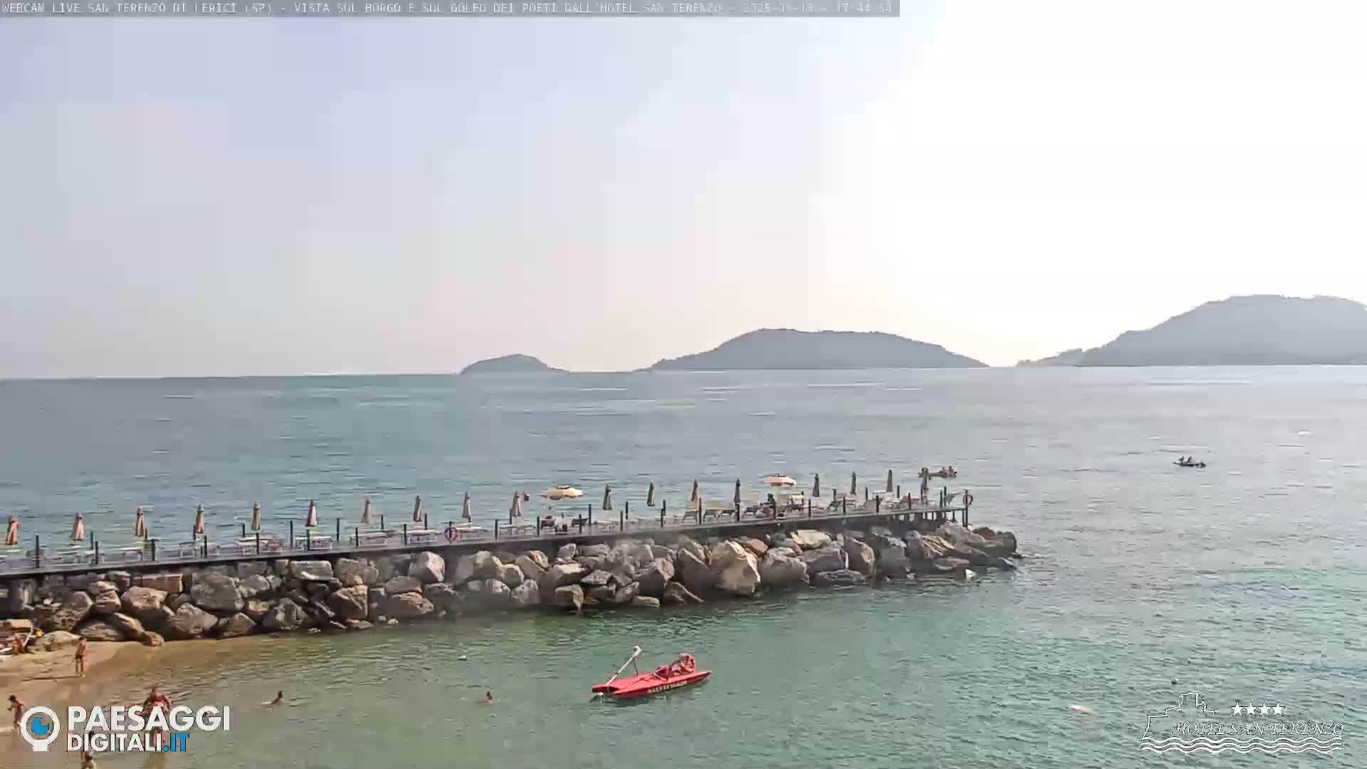 A long pier with umbrellas and people extends from a rocky shore into a calm sea, with distant hills visible under a hazy sky.