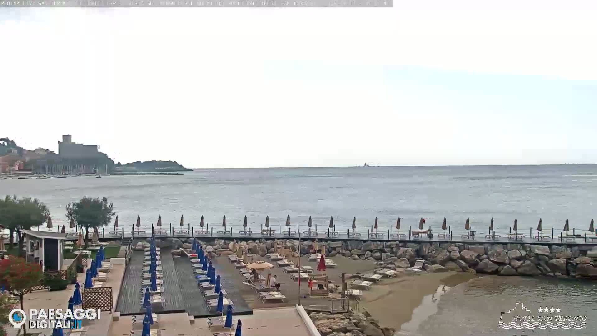 A beach with lounge chairs and umbrellas under an overcast sky overlooks a calm sea and a distant town with a castle.