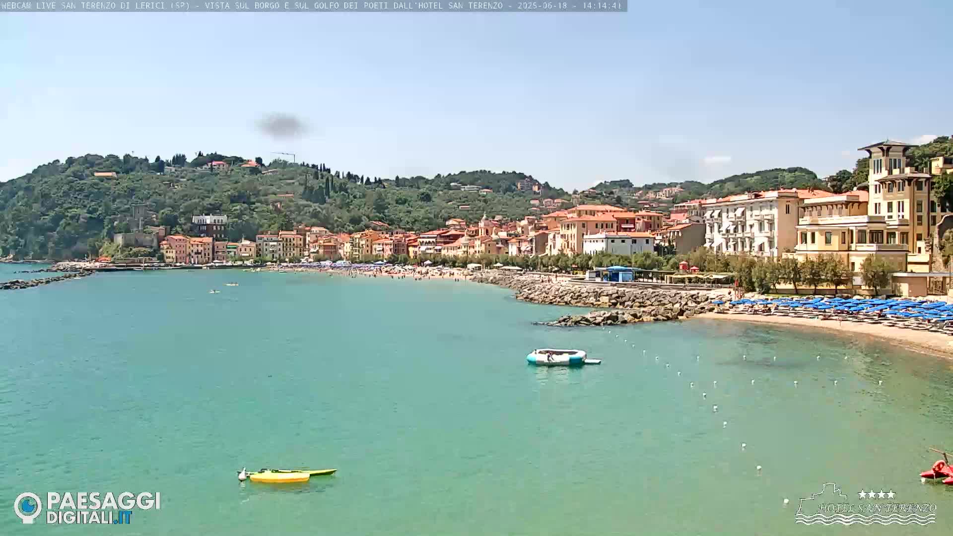 A picturesque coastal town nestled on a hillside, with a sandy beach and calm turquoise water under a clear blue sky.