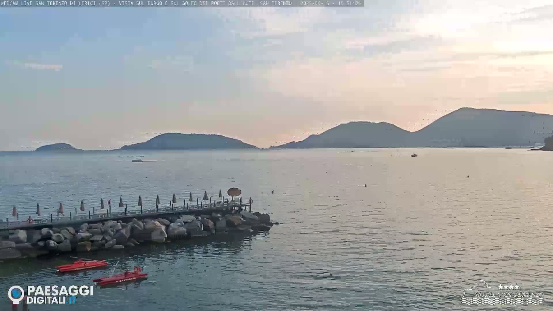 A calm sea with distant hills under a partly cloudy, pale sky, shows a rocky jetty with beach umbrellas and two red boats.