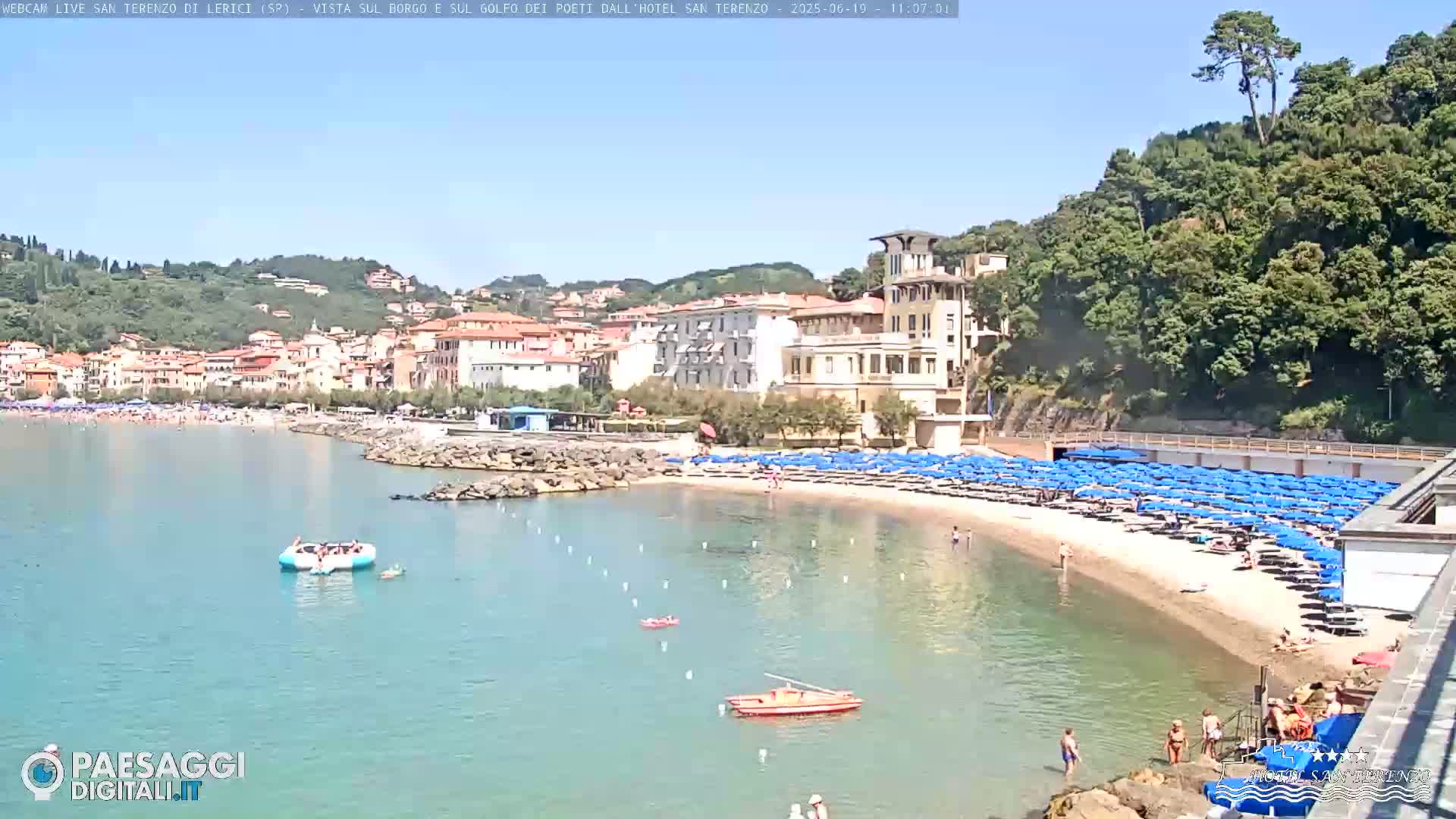 A sunny beach scene shows a sandy shoreline with numerous blue umbrellas, a calm turquoise bay, and a village nestled against green hills in the background.