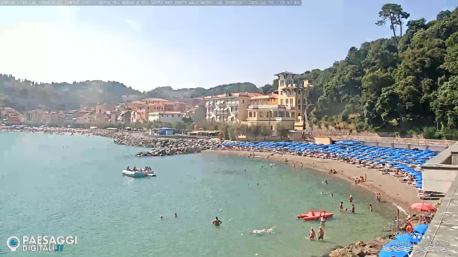 A sandy beach with numerous blue umbrellas and bathers is nestled in a calm bay, backed by a town and lush green hills under a clear, sunny sky.