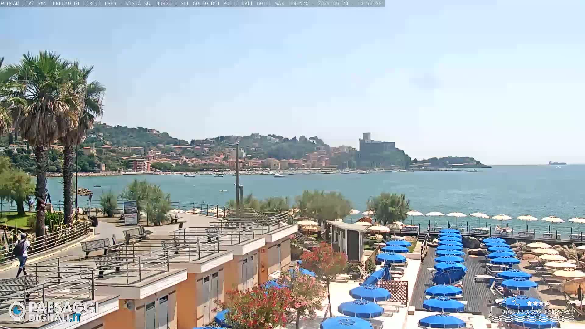 A sunny seaside town is seen from a high vantage point, overlooking a calm bay filled with boats and a beach lined with blue umbrellas.