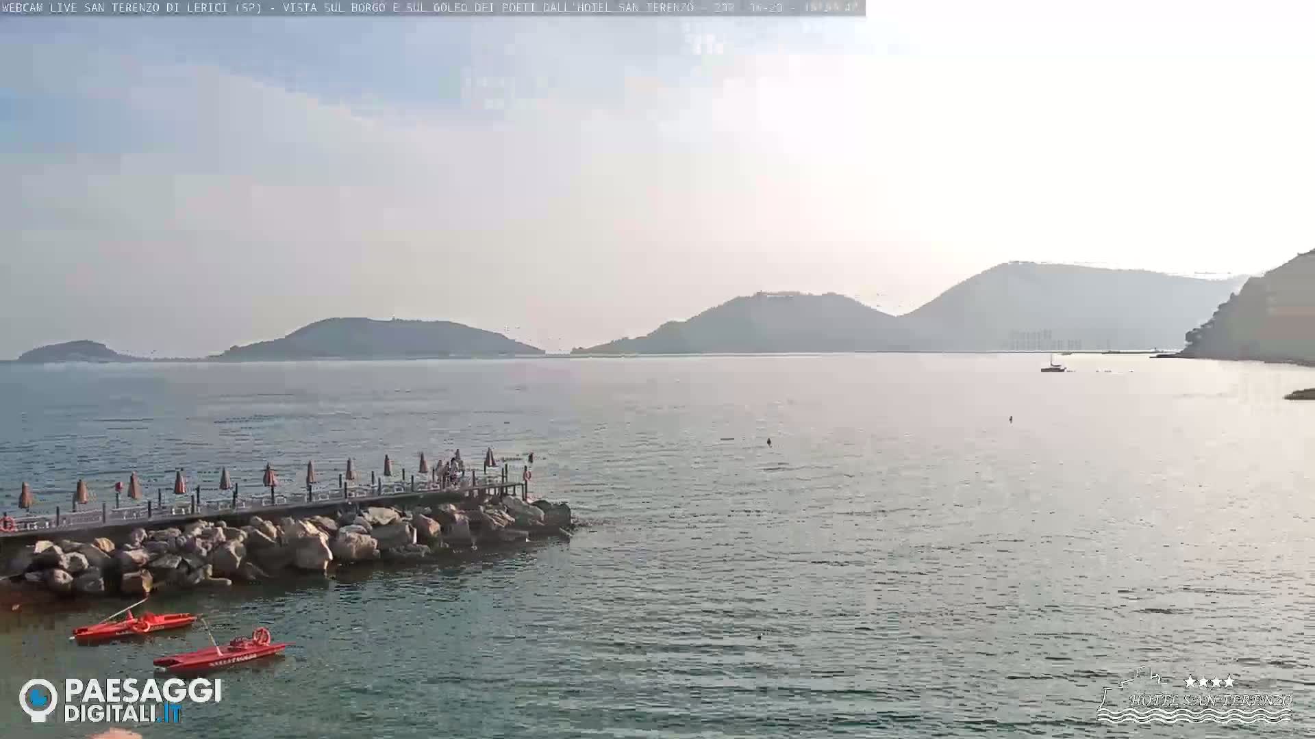 A hazy day reveals a calm body of water with a wooden pier, several beach umbrellas, and a few small boats, backed by distant hills.