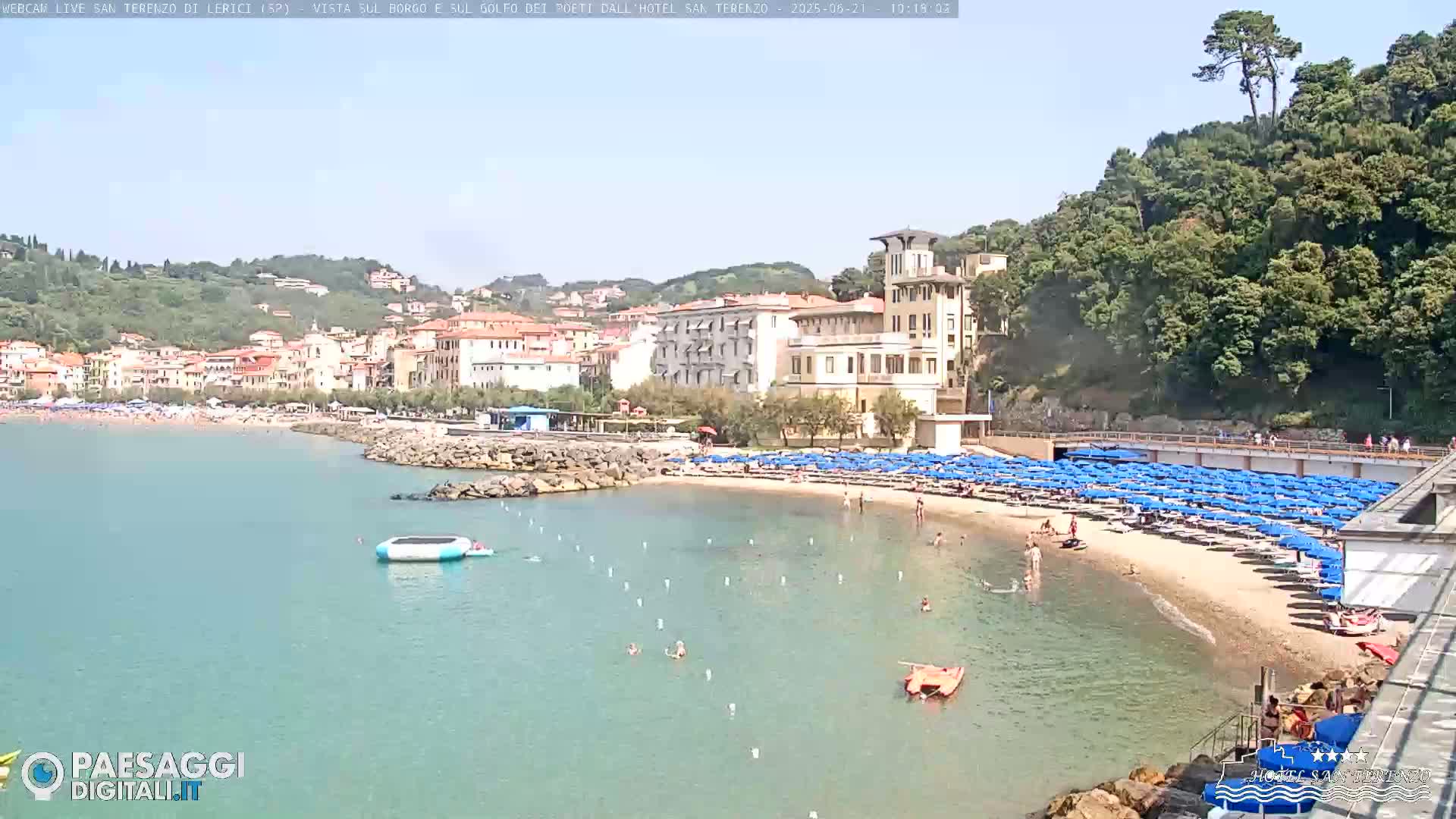 A sandy beach with numerous blue umbrellas and sunbathers is situated in front of a town nestled on a hillside, all under a bright, sunny sky.