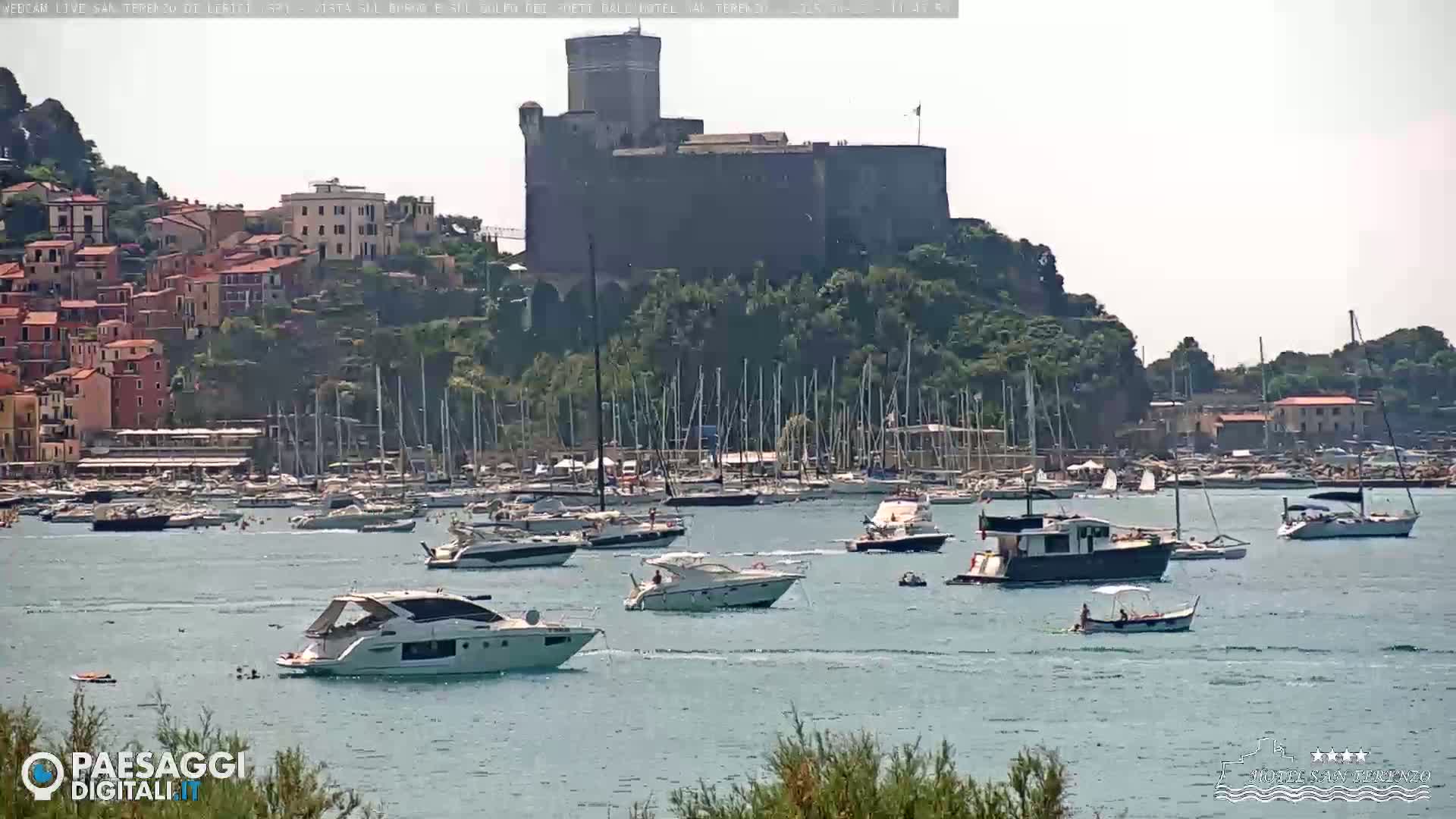 A stone castle sits atop a hill overlooking a harbor filled with numerous boats on a sunny day.