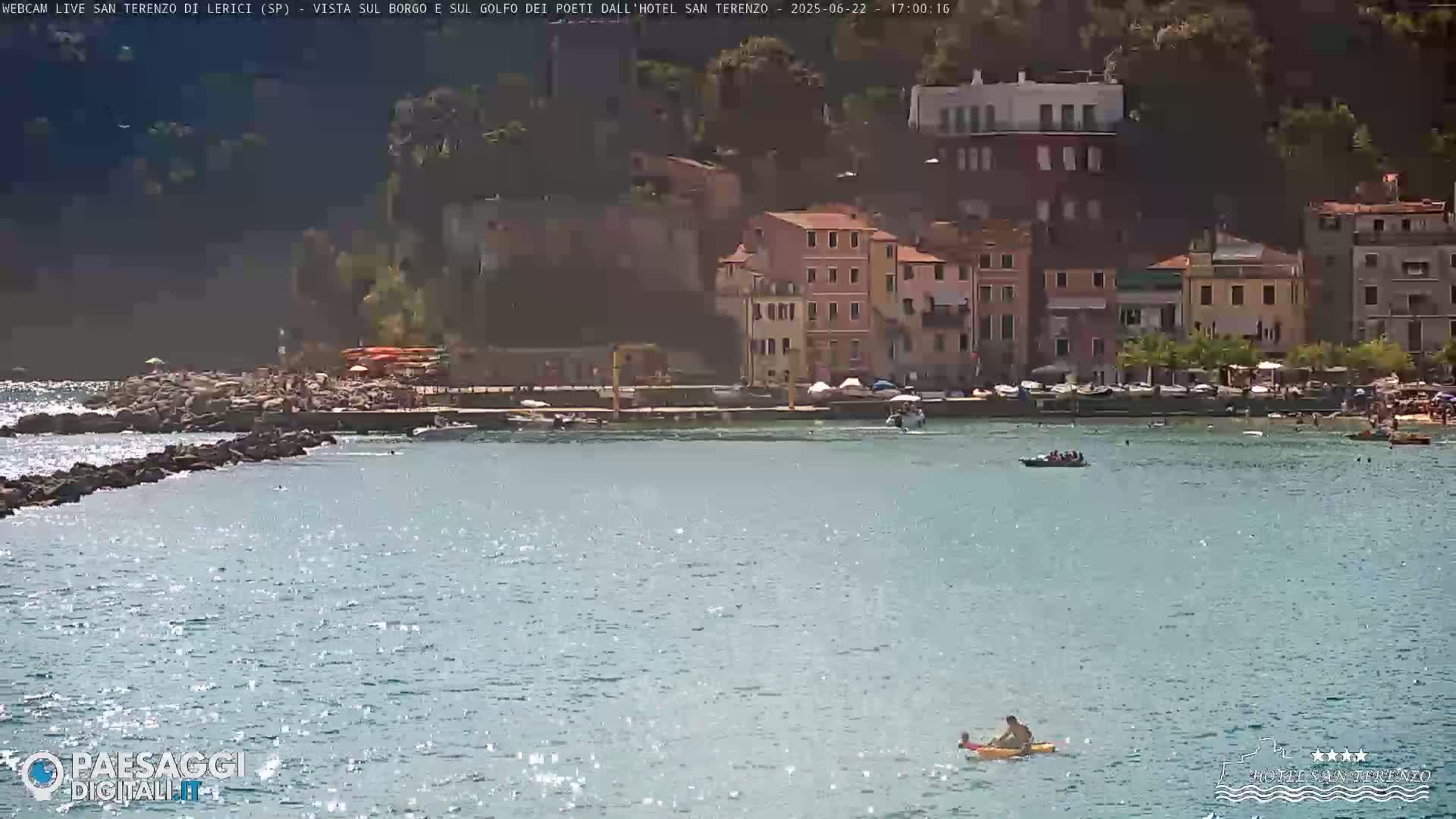 A sunny day shows a calm body of water with a few boats and people swimming near a rocky shore and a small village of colorful buildings on a hillside.