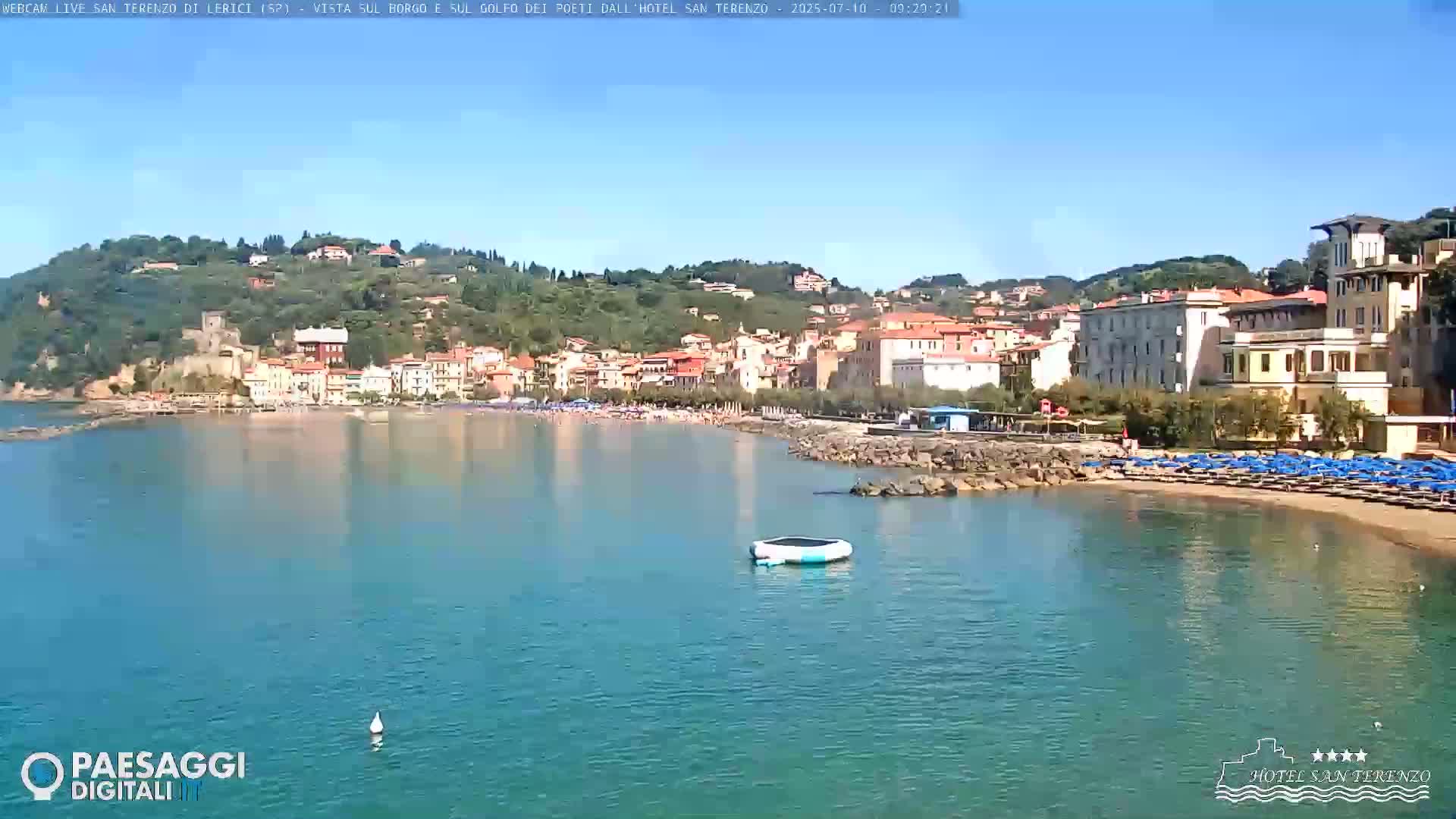 A coastal town with a sandy beach and calm, clear water is seen on a sunny day.