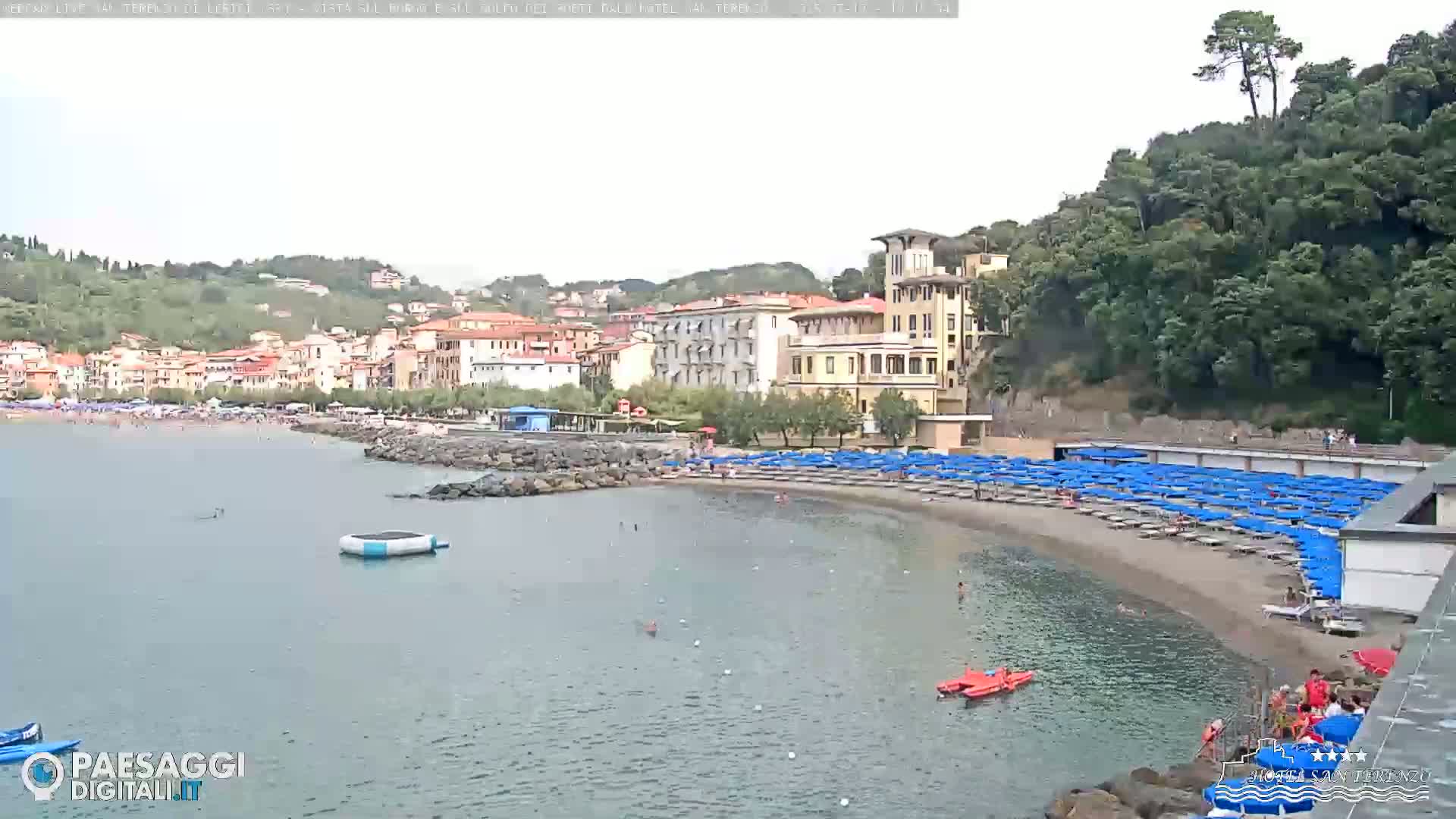 A calm bay with a sandy beach lined with blue umbrellas, backed by a village of buildings nestled against a hillside covered in lush green trees under an overcast sky.