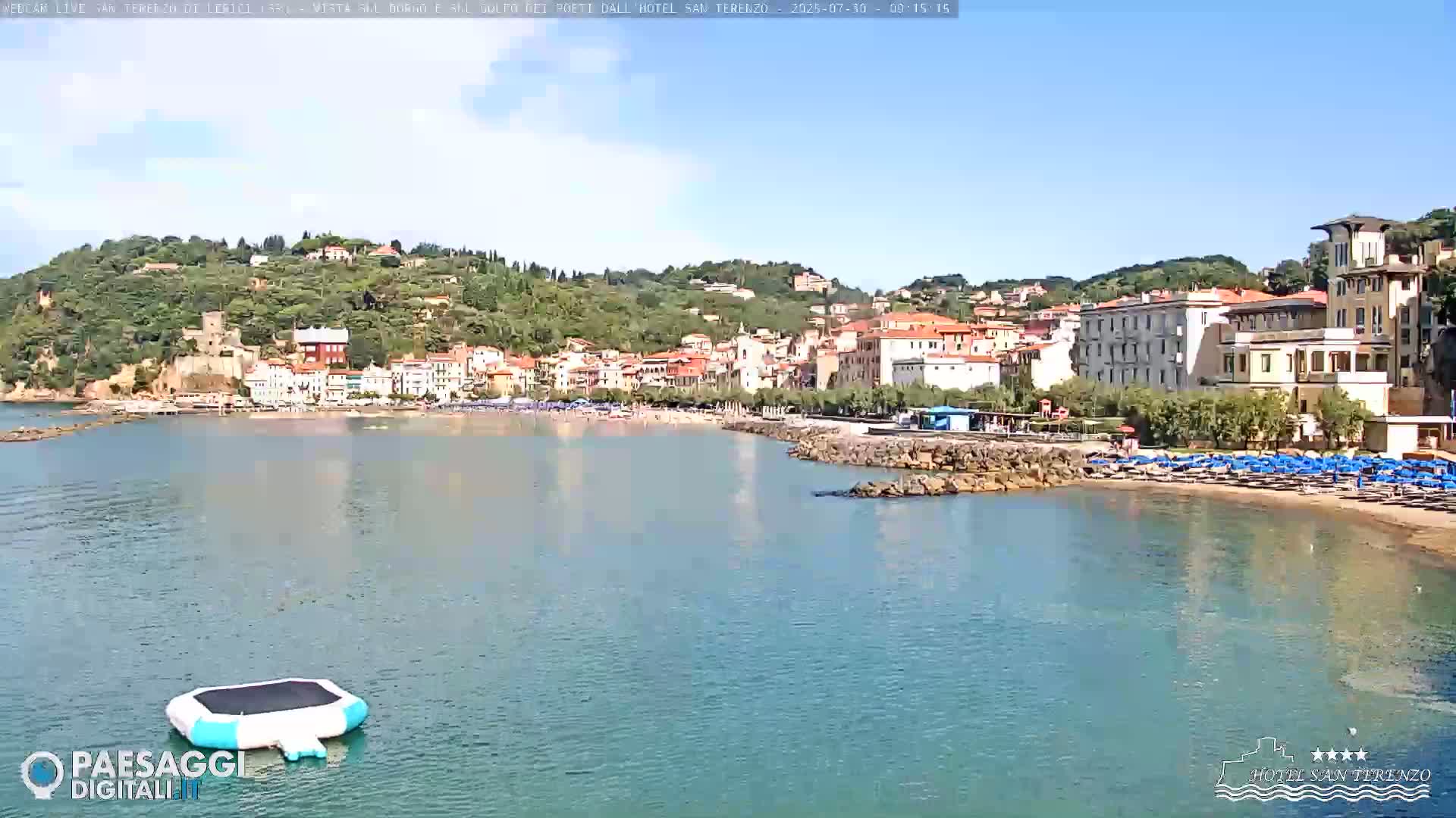 A calm bay with a sandy beach and a town built on a hillside under a clear sky.