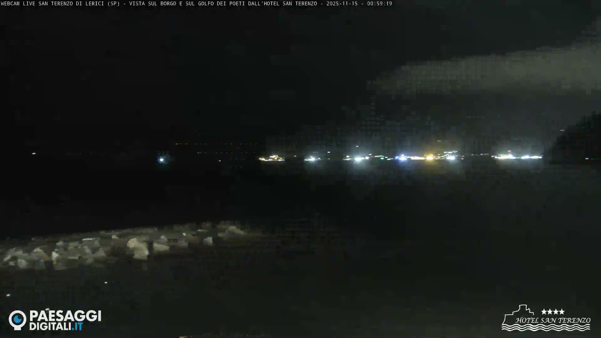 A dark nighttime view reveals a calm body of water reflecting the distant, illuminated outline of a coastal town or harbor under a clear sky, with subtle dark forms in the immediate foreground.