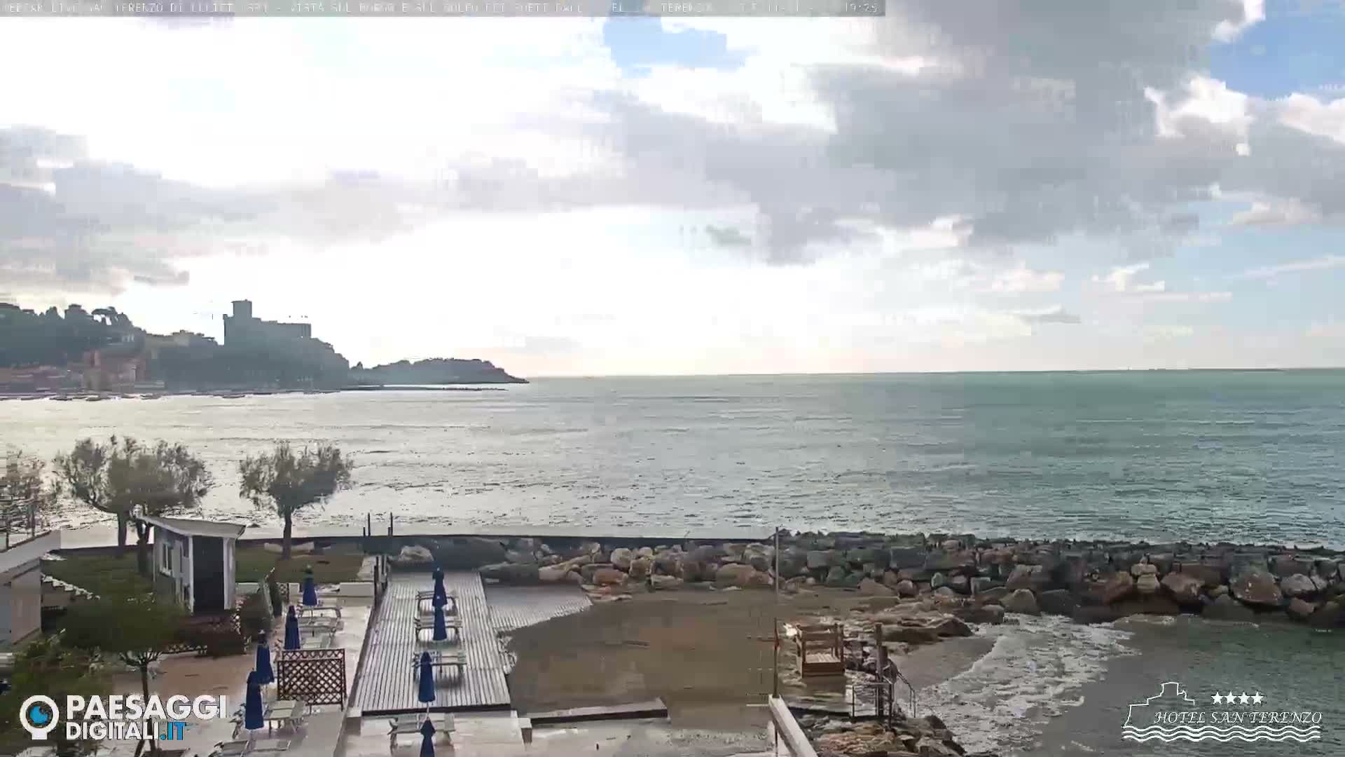 Under an overcast sky, a wide coastal shot reveals a beach foreground with wooden platforms and blue lounge chairs, bordered by a rocky shore and calm sea, extending to a distant town crowned by a castle on the left.
