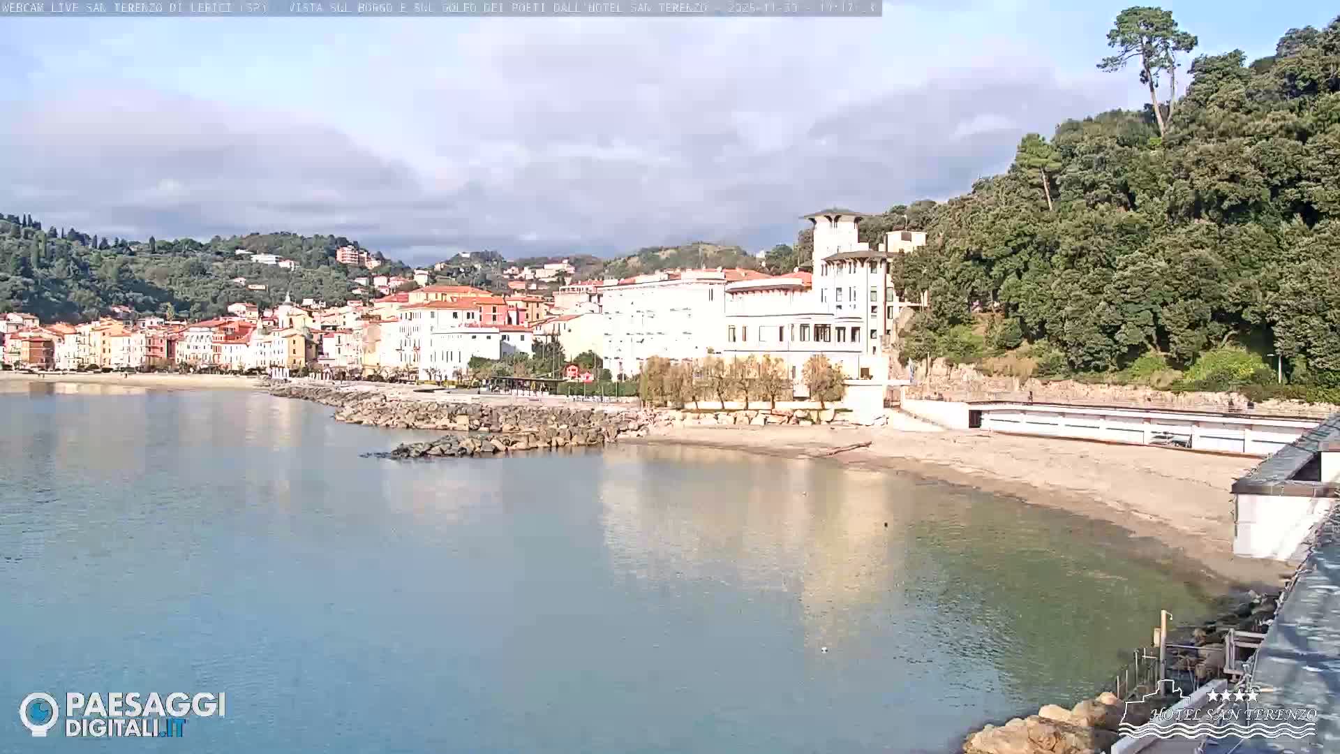 A picturesque coastal town with colorful buildings and a prominent white hotel lines a calm bay featuring a sandy beach and rock barriers, all set against a backdrop of densely forested hills under a partly cloudy sky.