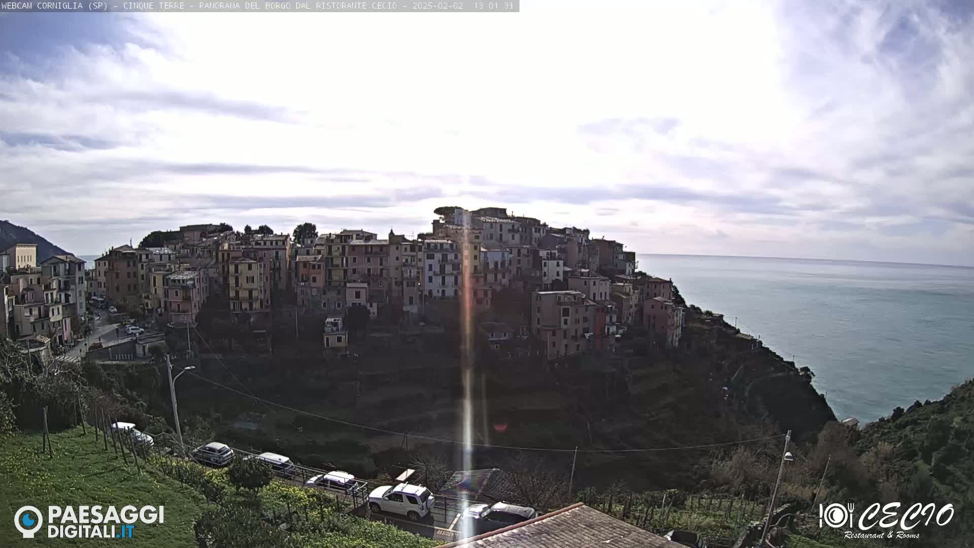 A coastal village of closely clustered buildings sits atop a cliff overlooking a calm sea under a mostly cloudy sky.