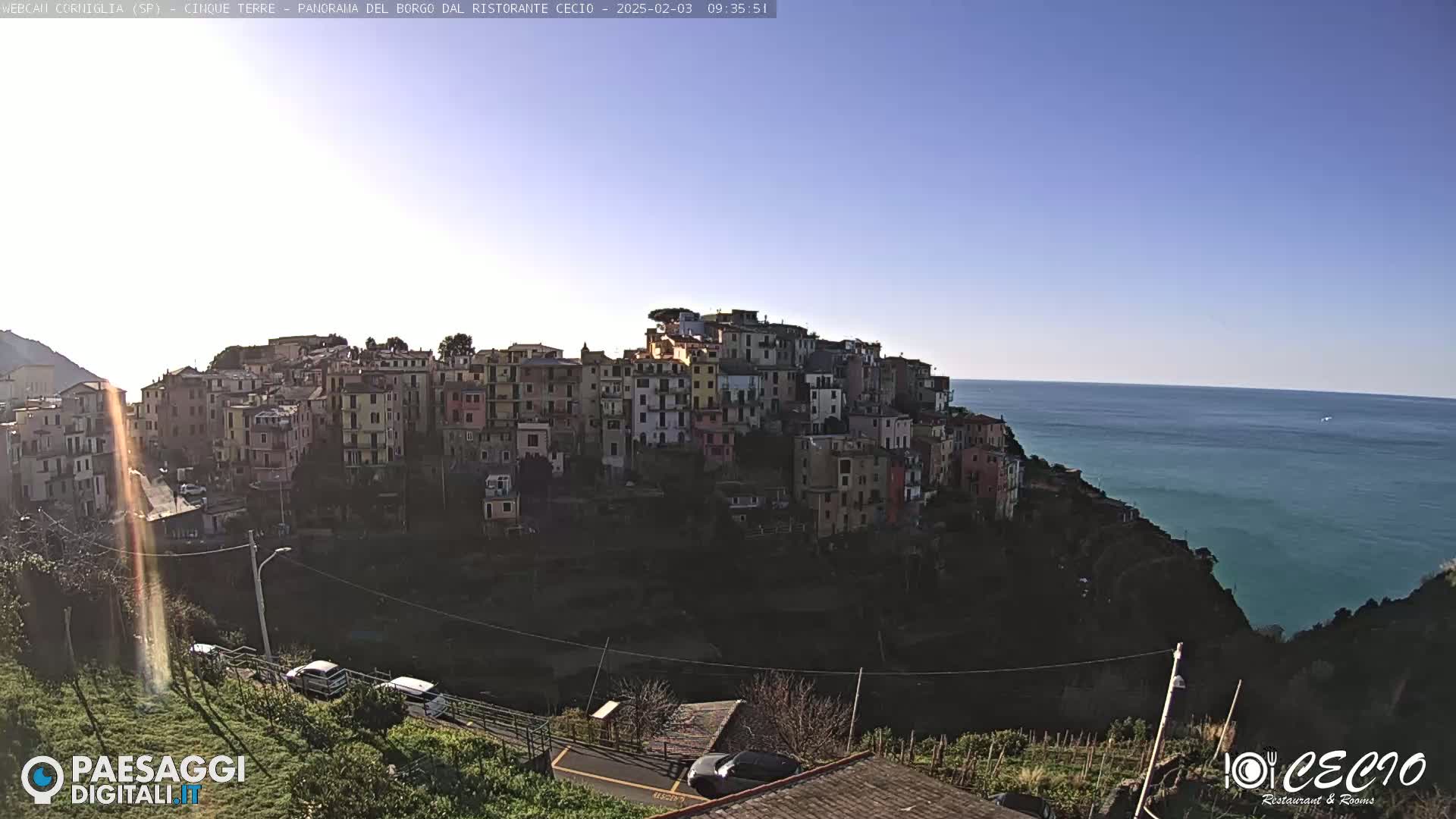 A colorful village clings to a hillside overlooking a calm, blue sea under a clear, bright sky.