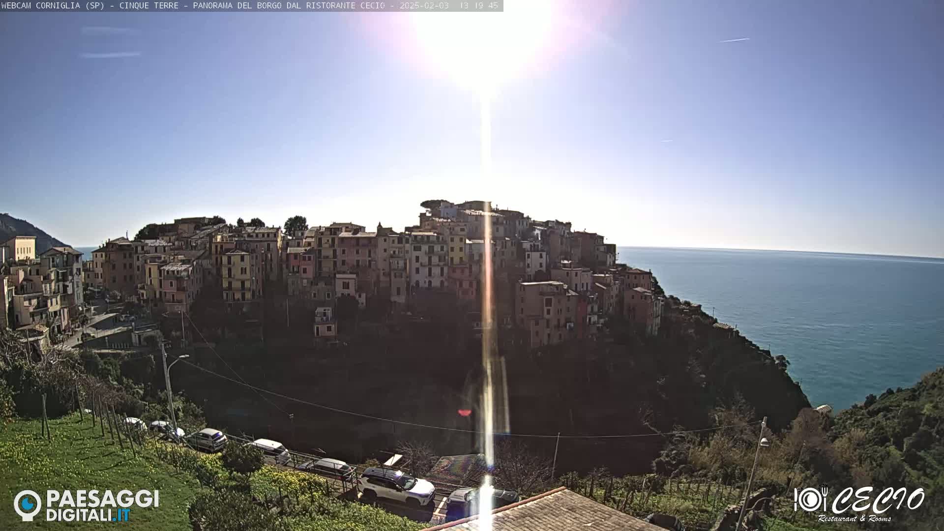 A sunlit coastal village of colorful buildings clings to a hillside overlooking a calm sea under a clear sky.