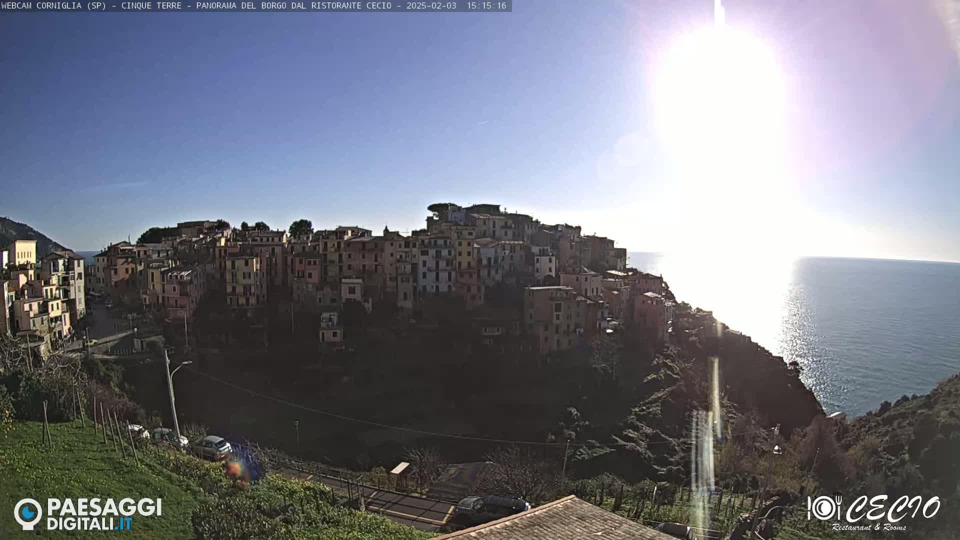 A sunny day reveals a coastal village of closely clustered buildings perched on a hillside overlooking a calm sea.