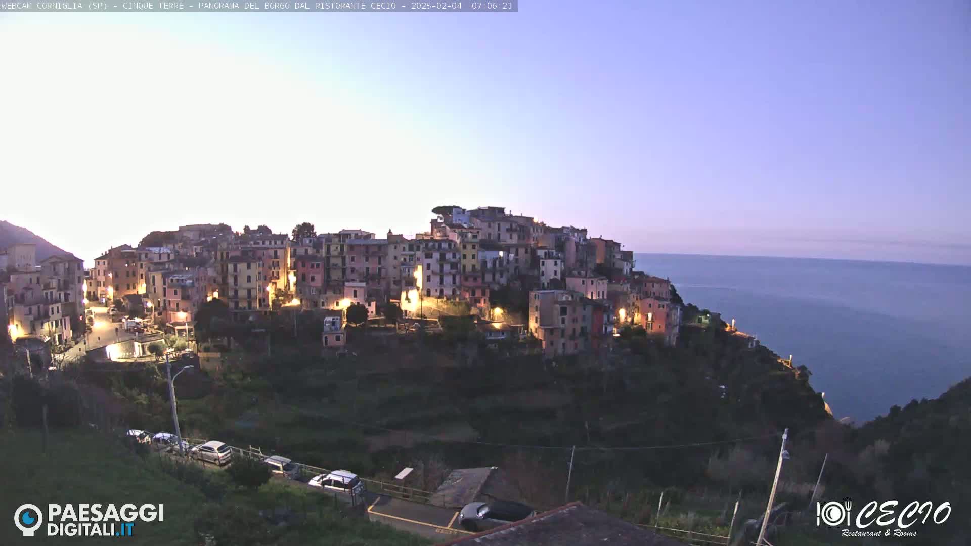 A brightly lit village clings to a hillside overlooking a calm, dark sea under a twilight sky.