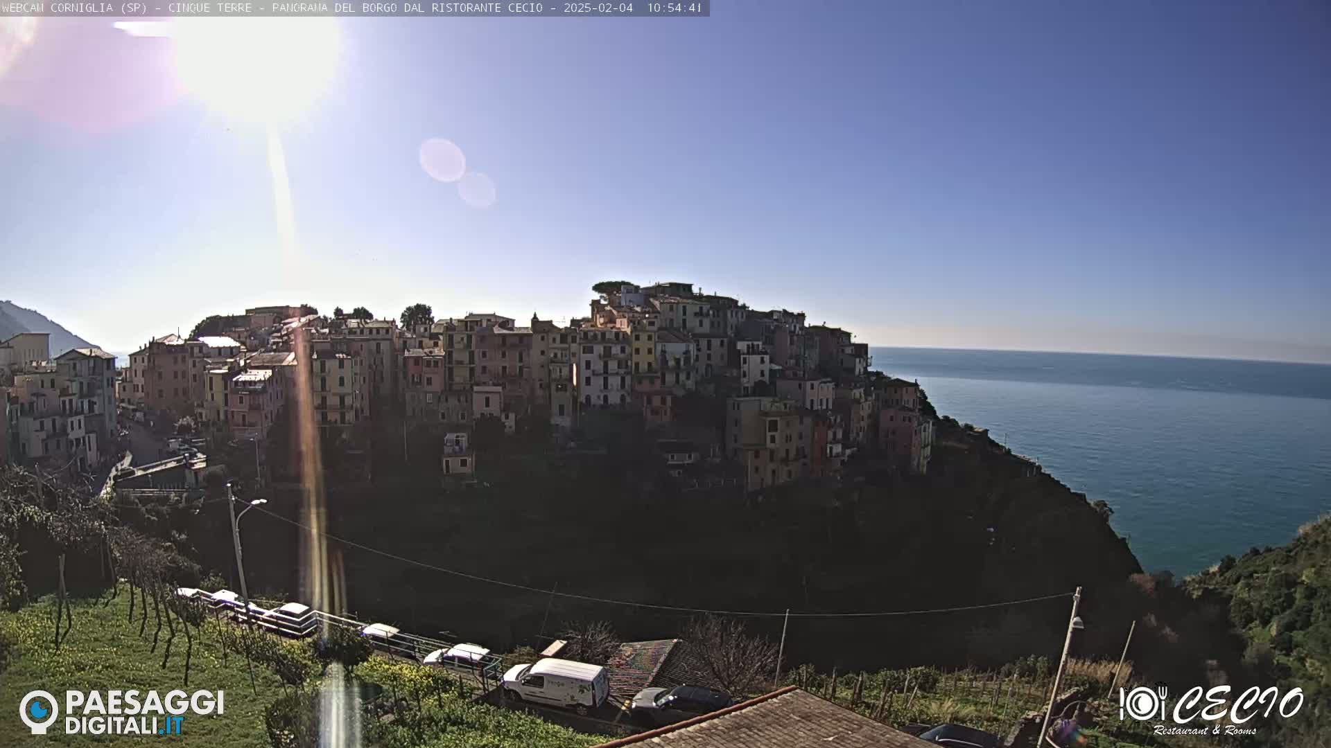 A brightly lit coastal town of closely clustered buildings sits atop a cliff overlooking a calm blue sea under a clear sunny sky.
