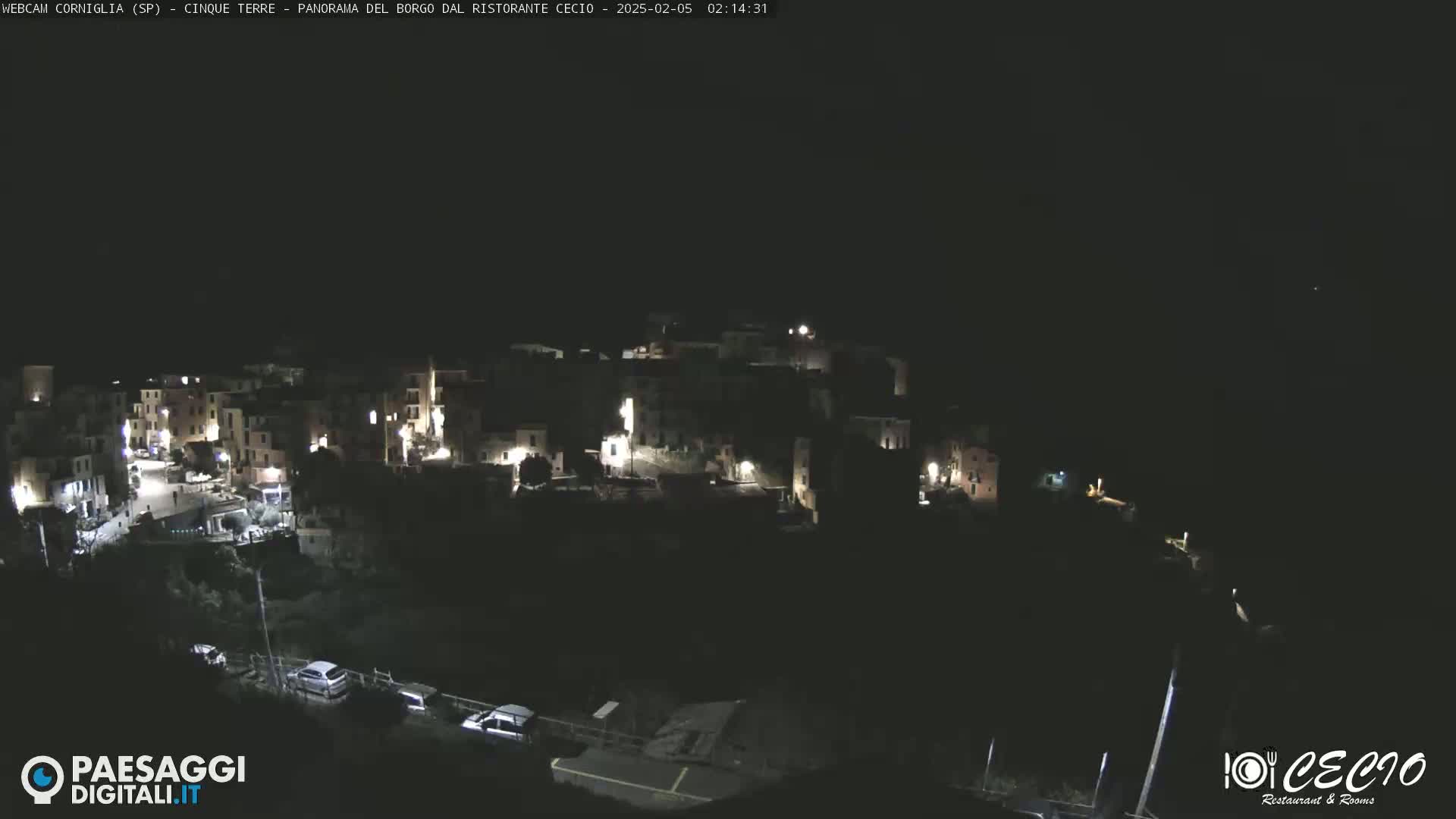 A nighttime, aerial view of a small, dark village with sparsely lit buildings and a few cars parked near the bottom.