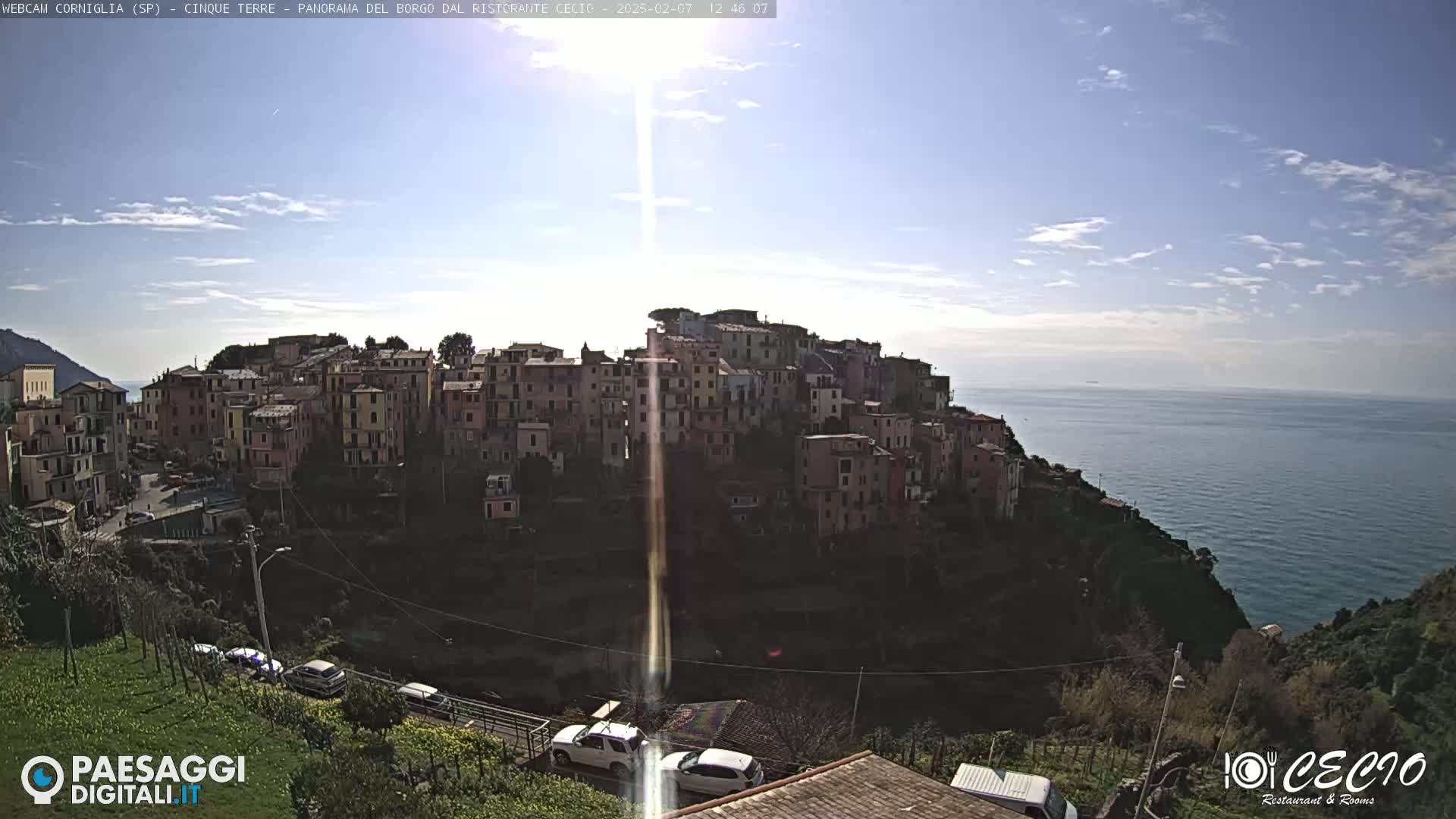 A sunlit coastal village of colorful buildings clings to a hillside overlooking a calm sea under a mostly clear sky.