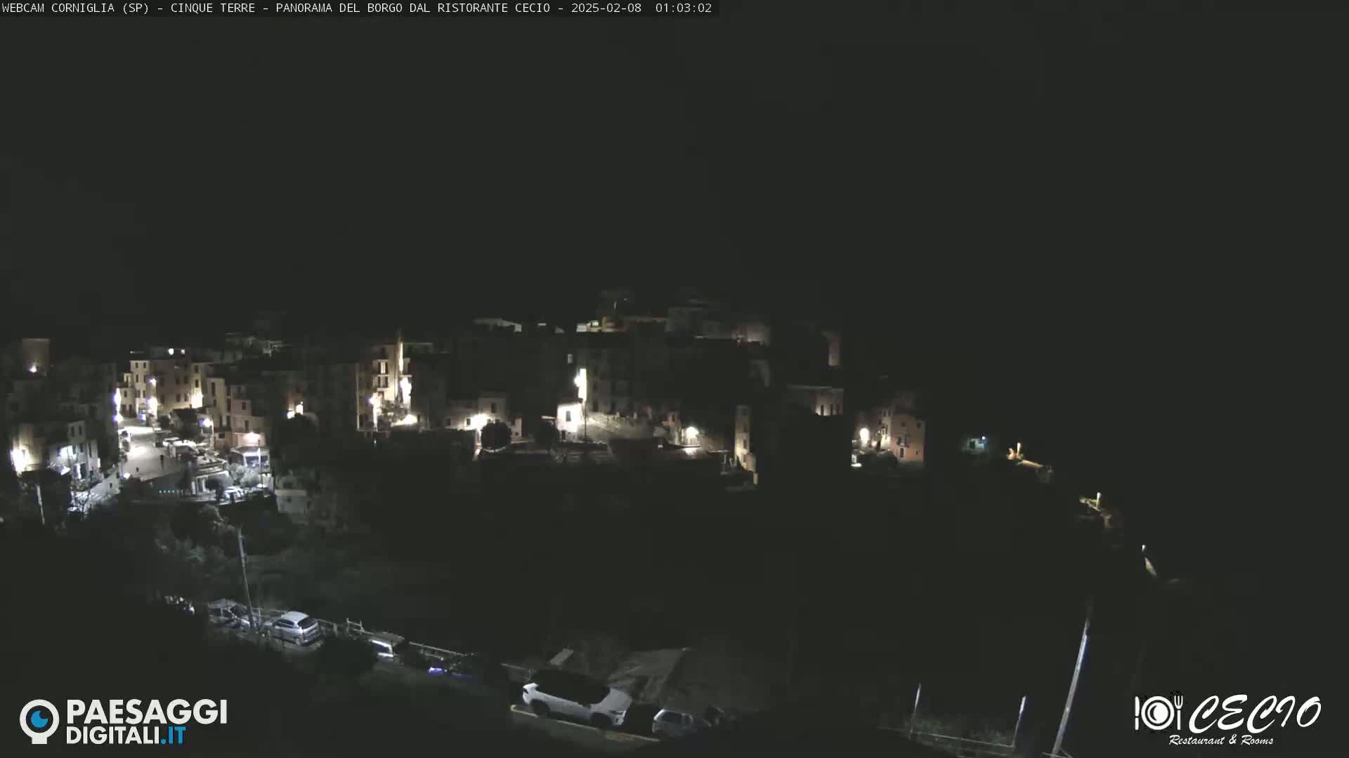 A nighttime, high-angle view shows a small, dimly lit village nestled against a dark hillside.