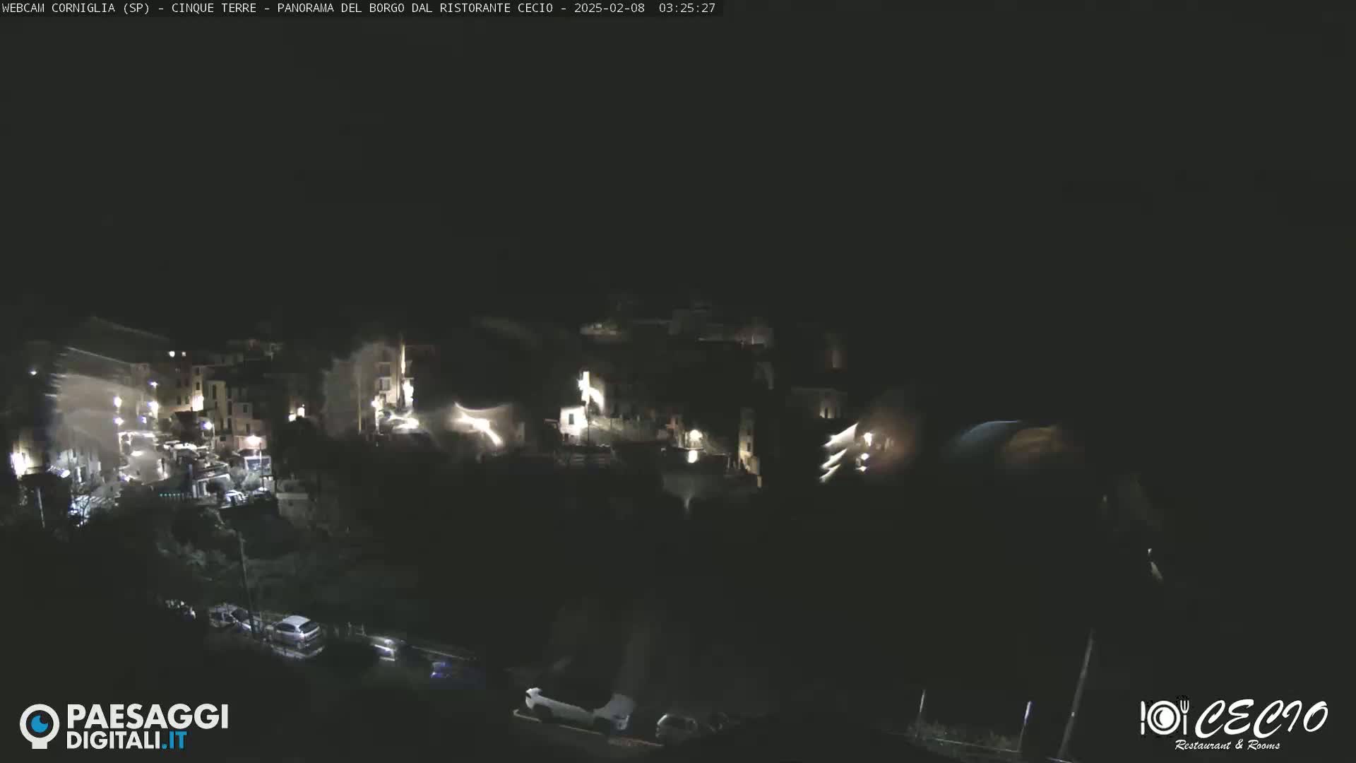 A nighttime, high-angle view shows a small, dimly lit village nestled into a dark hillside with a few vehicles visible on a road below.