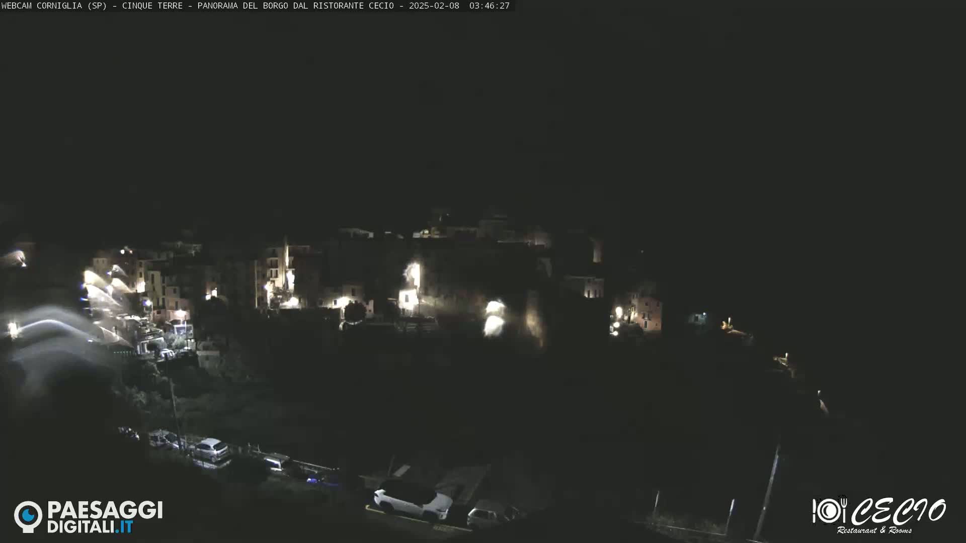 A nighttime, high-angle view of a small, dark village with scattered lights, a few cars parked below, and a mostly clear sky.