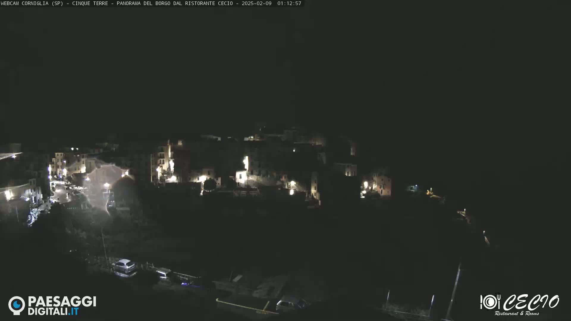 A nighttime, high-angle view shows a small, dark village with sparsely lit buildings, and a few cars are parked near the bottom of a dimly lit road.