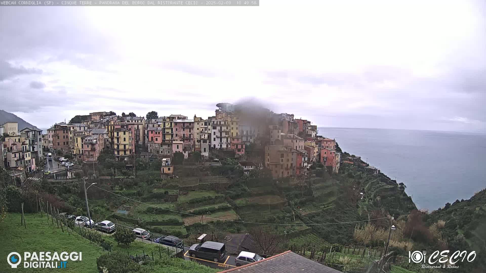 A coastal town of colorful buildings clings to a hillside overlooking a calm sea under an overcast sky.