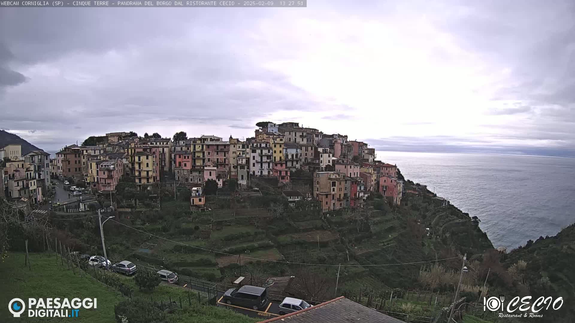 A hillside village of colorful buildings overlooks a calm sea under an overcast sky.