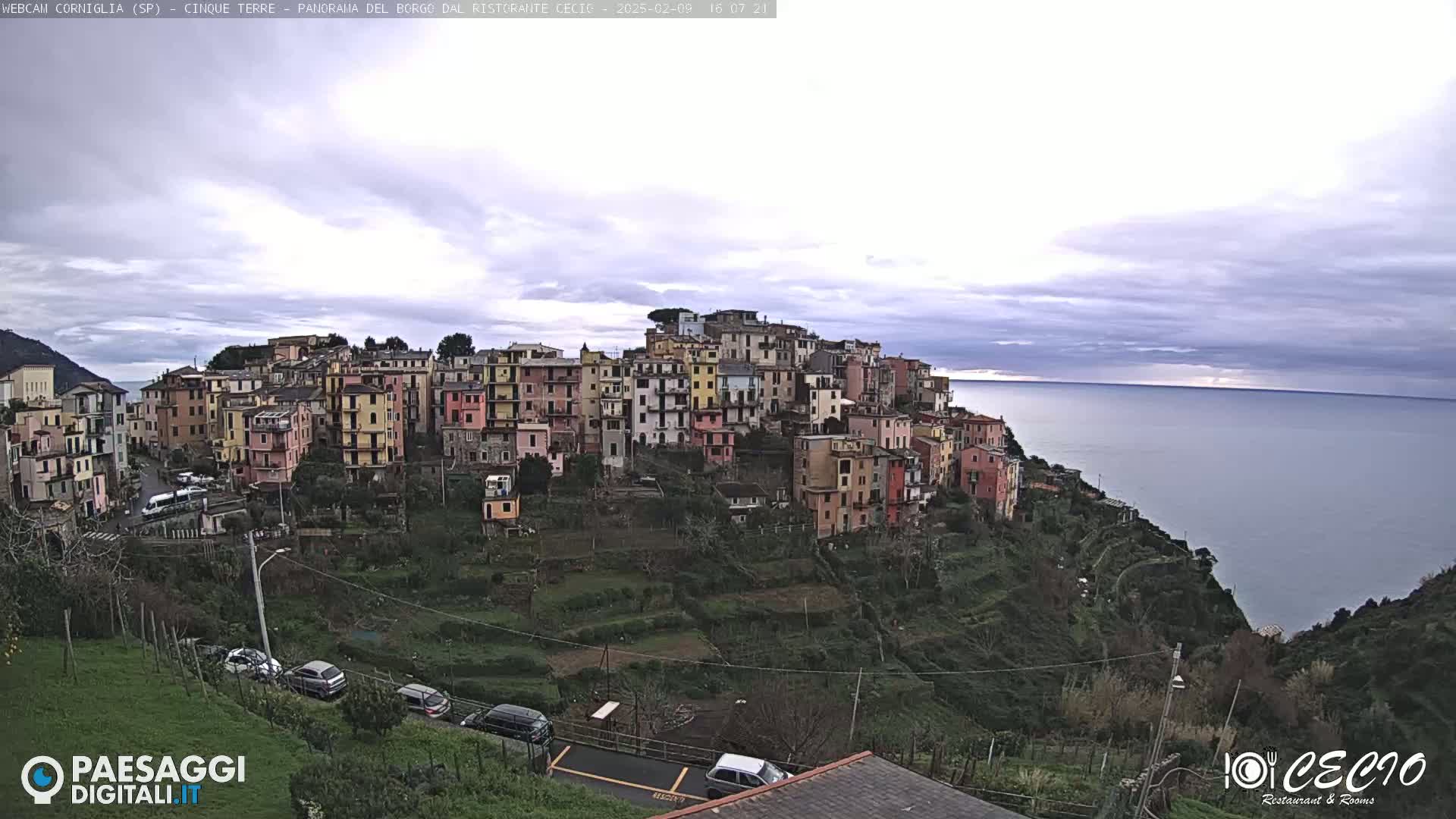 A hillside village of colorful buildings overlooks a calm ocean under a cloudy sky.