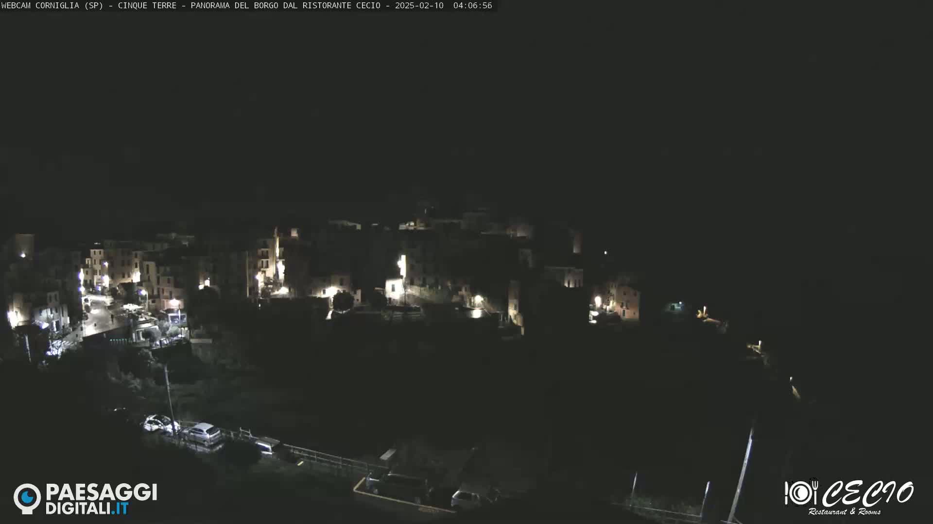 A nighttime, aerial view of a small, dark village with scattered lights, a few cars parked near a dark body of water.