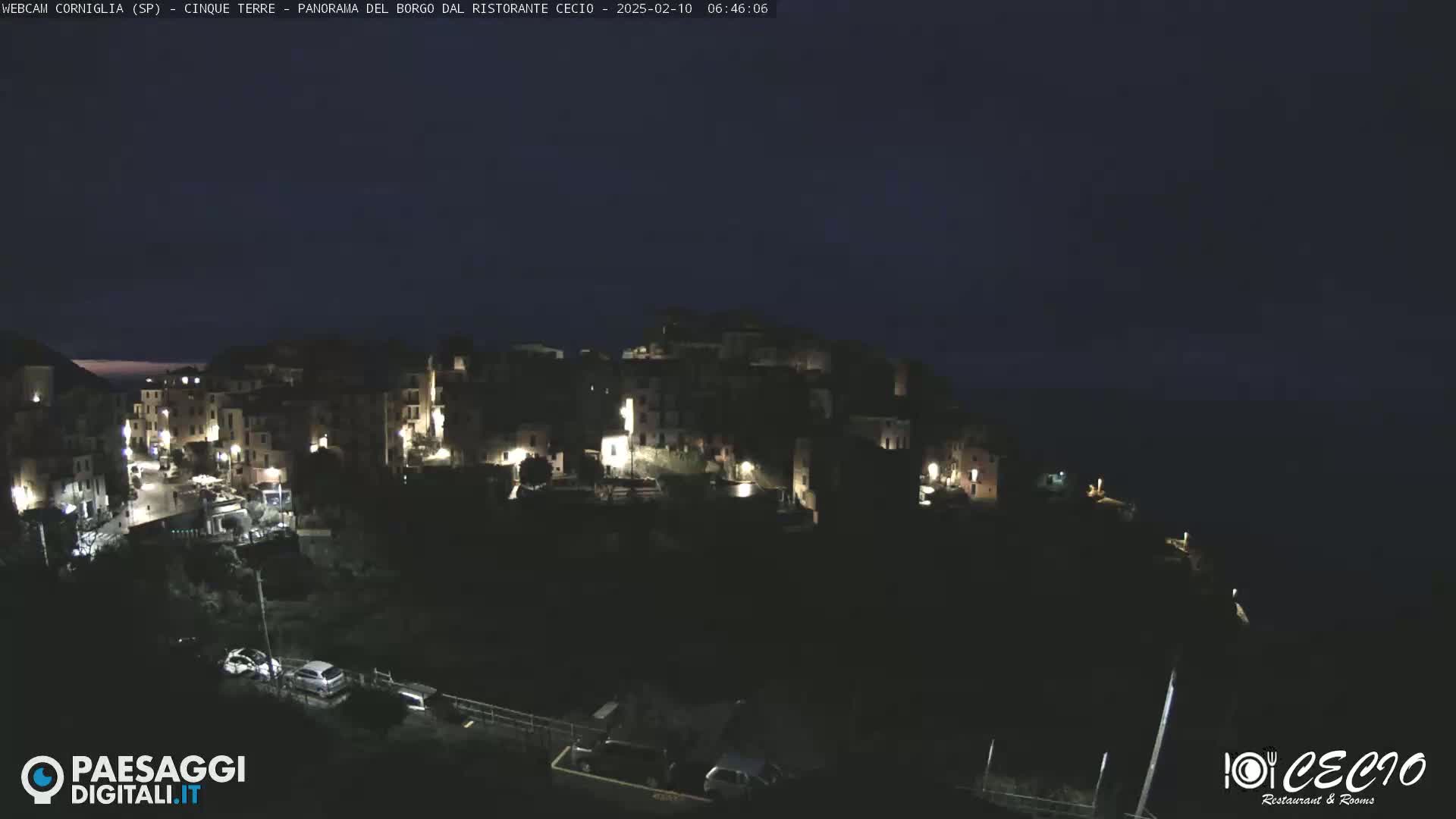 A nighttime view of a small, illuminated town nestled on a cliffside overlooking a dark body of water under a clear, dark sky.
