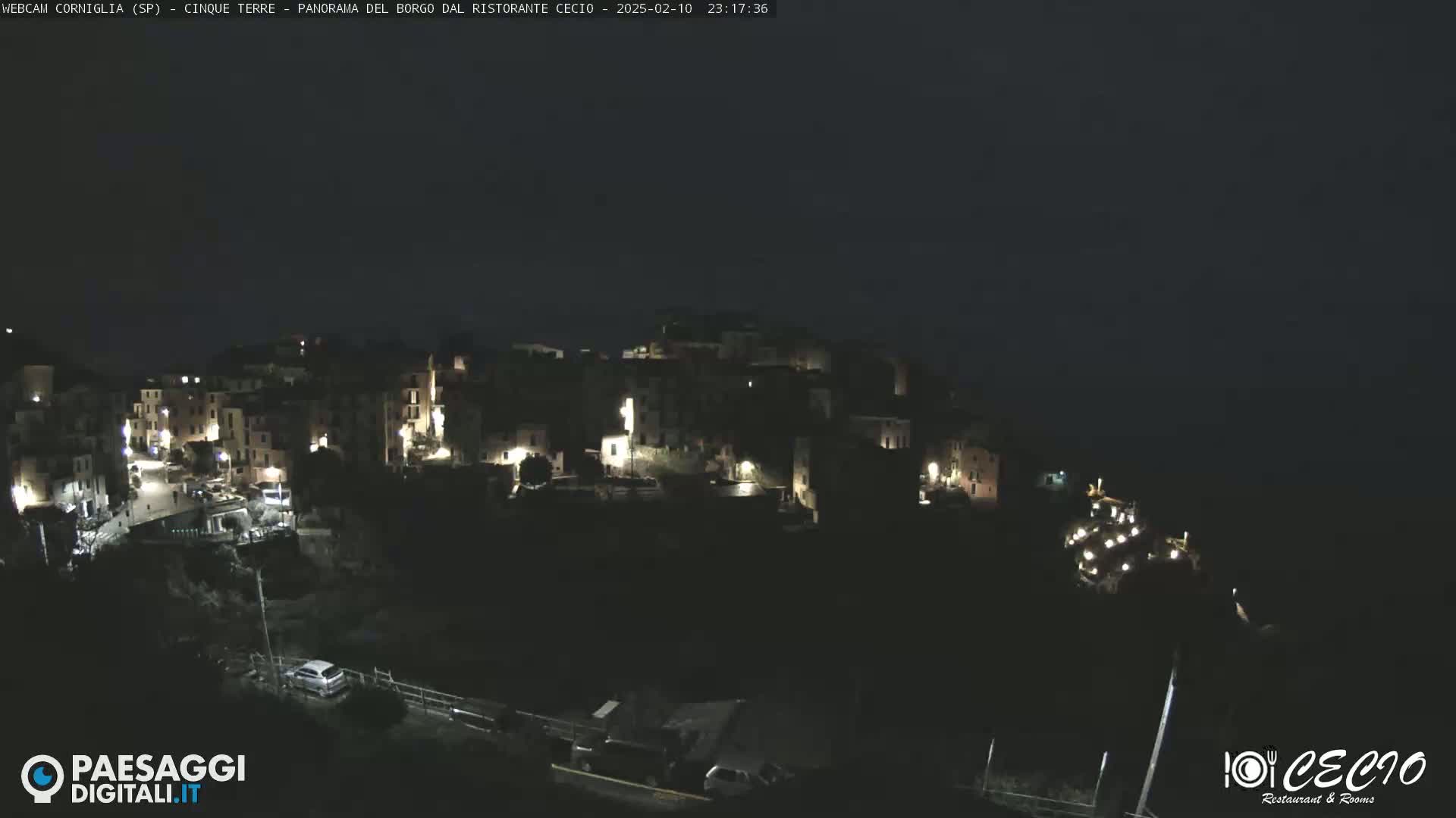 A nighttime, high-angle view of a small, hilly town with illuminated buildings, partially obscured by darkness.