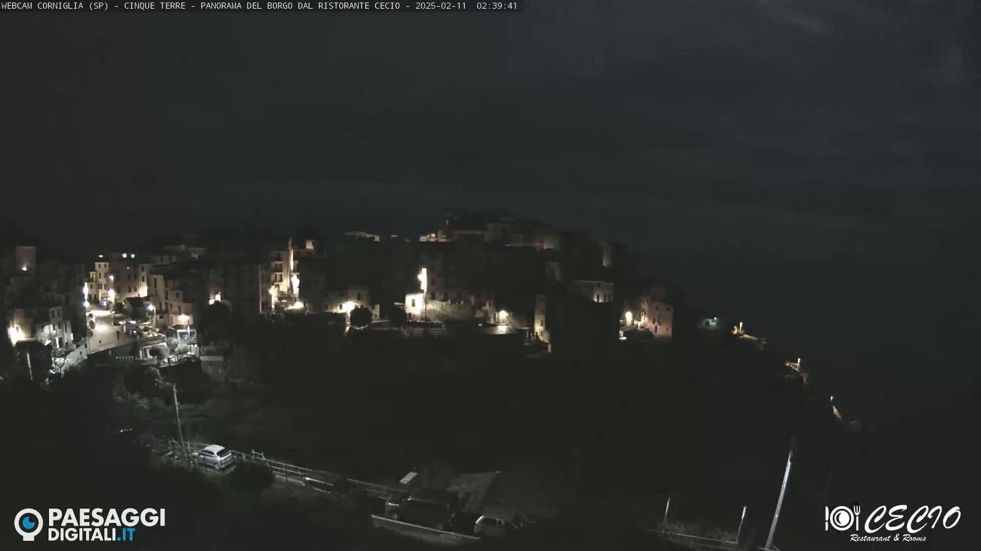 A nighttime, high-angle view of a small, hilltop town with dimly lit buildings, overlooking a dark body of water under a cloudy sky.