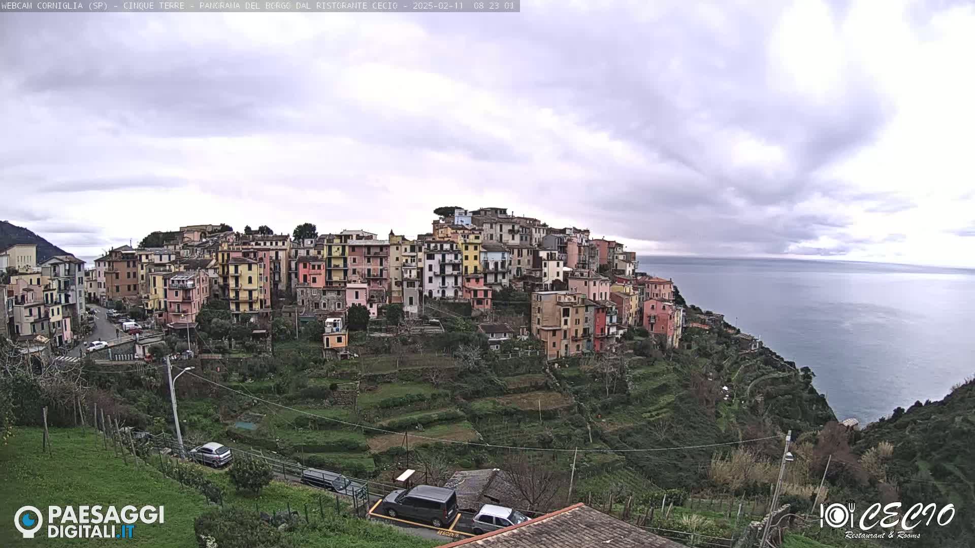 A hillside village of colorful buildings overlooks a calm sea under a mostly cloudy sky.